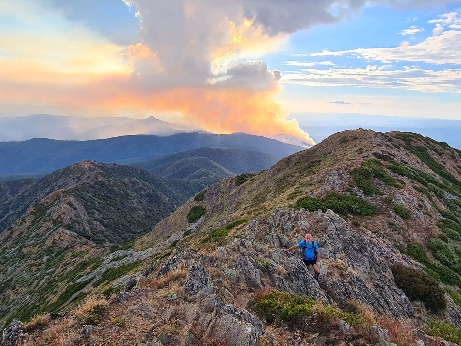 登山 桌布 壁紙 — 一名遠足者勝利地站在崎嶇的山脊上，熾熱的夕陽點燃了遠處山峰背後的雲彩。