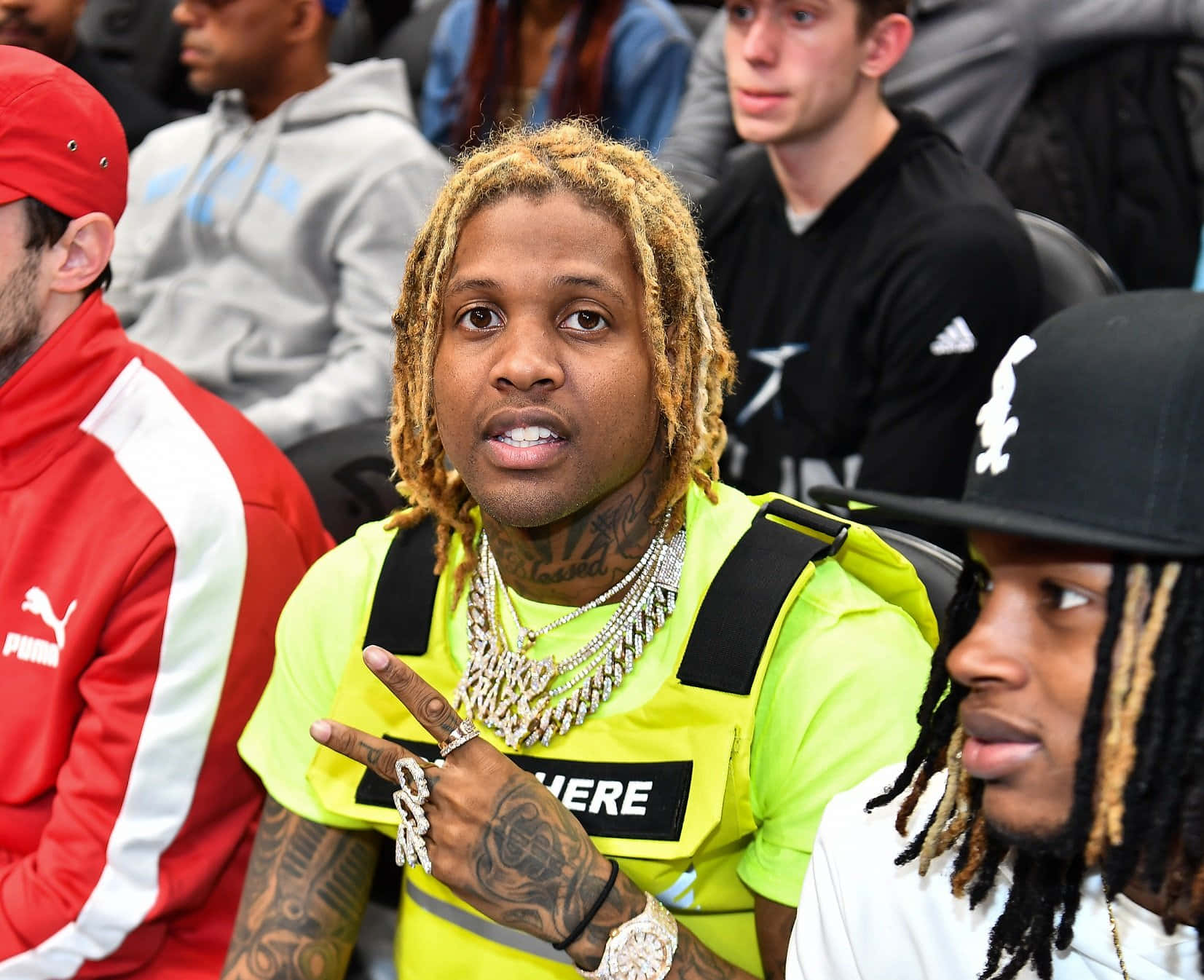 Lil Durk 桌布 壁紙 — 一名金色辮子及佩戴重型珠寶的男子坐在擁擠的場館中比出和平手勢。