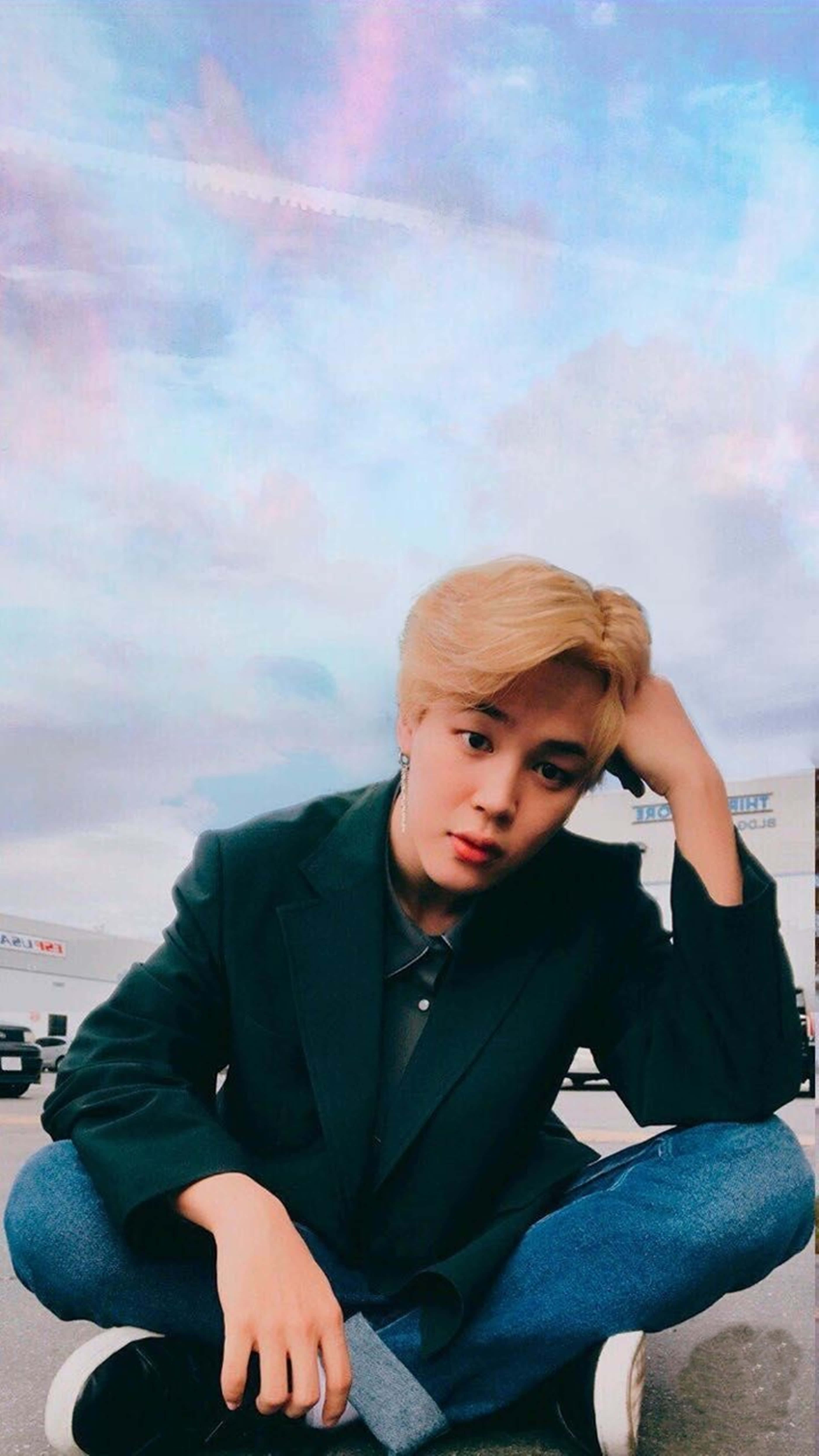 Jimin Bts Cute 桌布 壁紙 — 一名金髮年輕男子盤腿坐在人行道上，上方是帶有彩虹色雲朵的鮮豔粉彩天空。