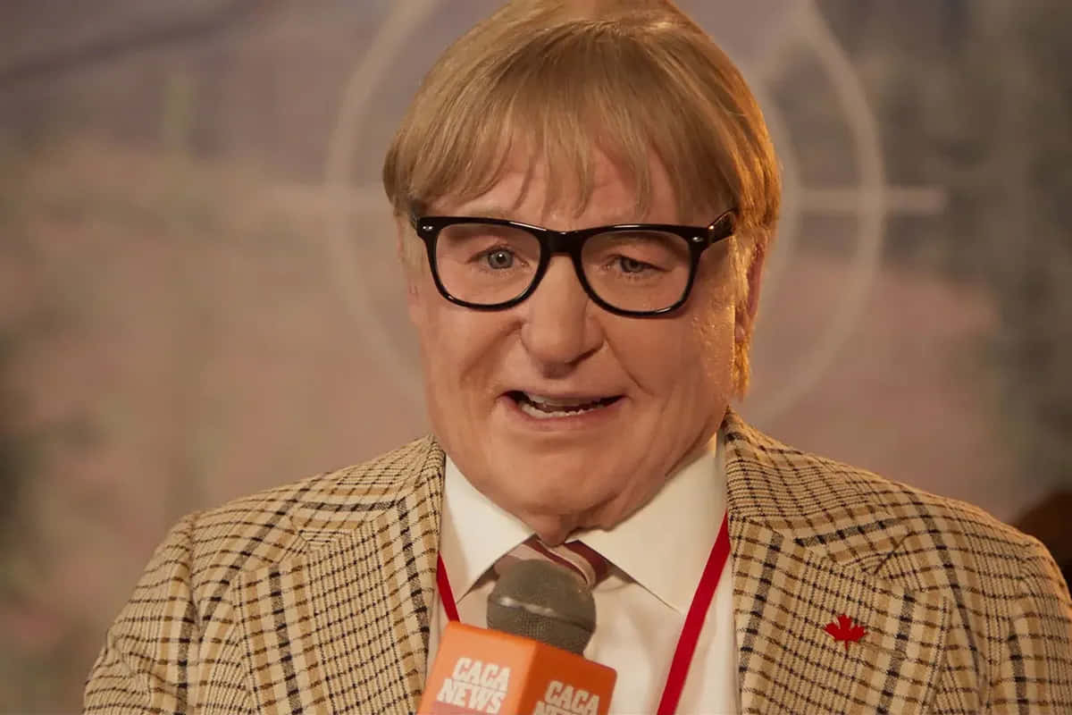 Mike Myers 桌布 壁紙 — 一名金髮戴黑框眼鏡的男子，在模糊背景前微笑，手持印有「CACA NEWS」的麥克風。