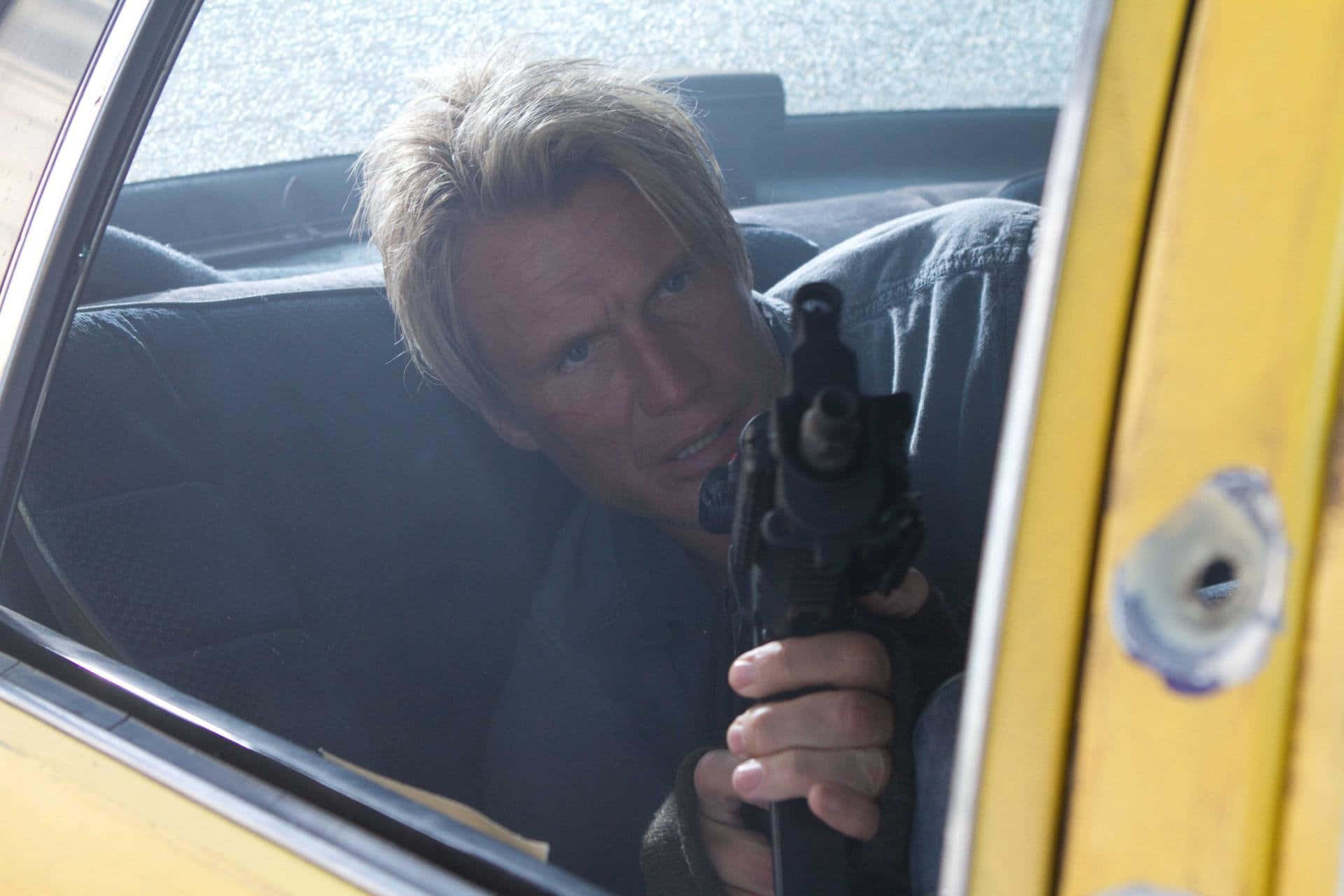 Dolph Lundgren 桌布 壁紙 — 一名金髮男子從黃色車窗探出頭來，手持槍枝，車門上可見彈孔。