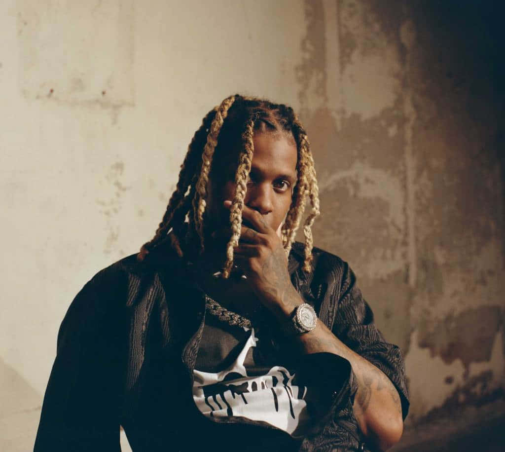 Lil Durk 桌布 壁紙 — 一名金髮辮子、有刺青的男子，在風化的牆壁前沉思擺姿勢，手靠近嘴邊，身穿黑色外套和骷髏圖案T恤。