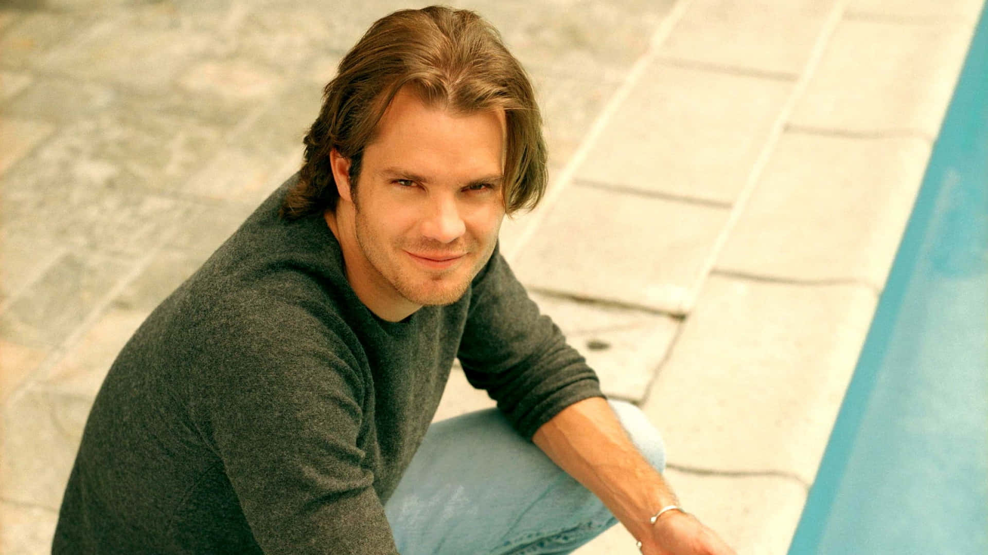 timothy olyphant 桌布 壁紙 — 一名長棕髮的男子在陽光普照的日子蹲在游泳池旁微笑。