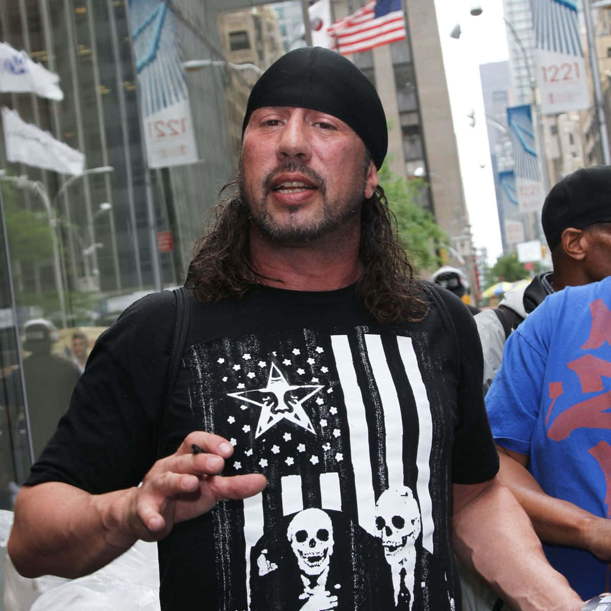 WWE 桌布 壁紙 — 一名長髮戴黑色毛帽的男子，在繁忙的城市街道上穿著骷髏主題的美國國旗襯衫並做手勢。
