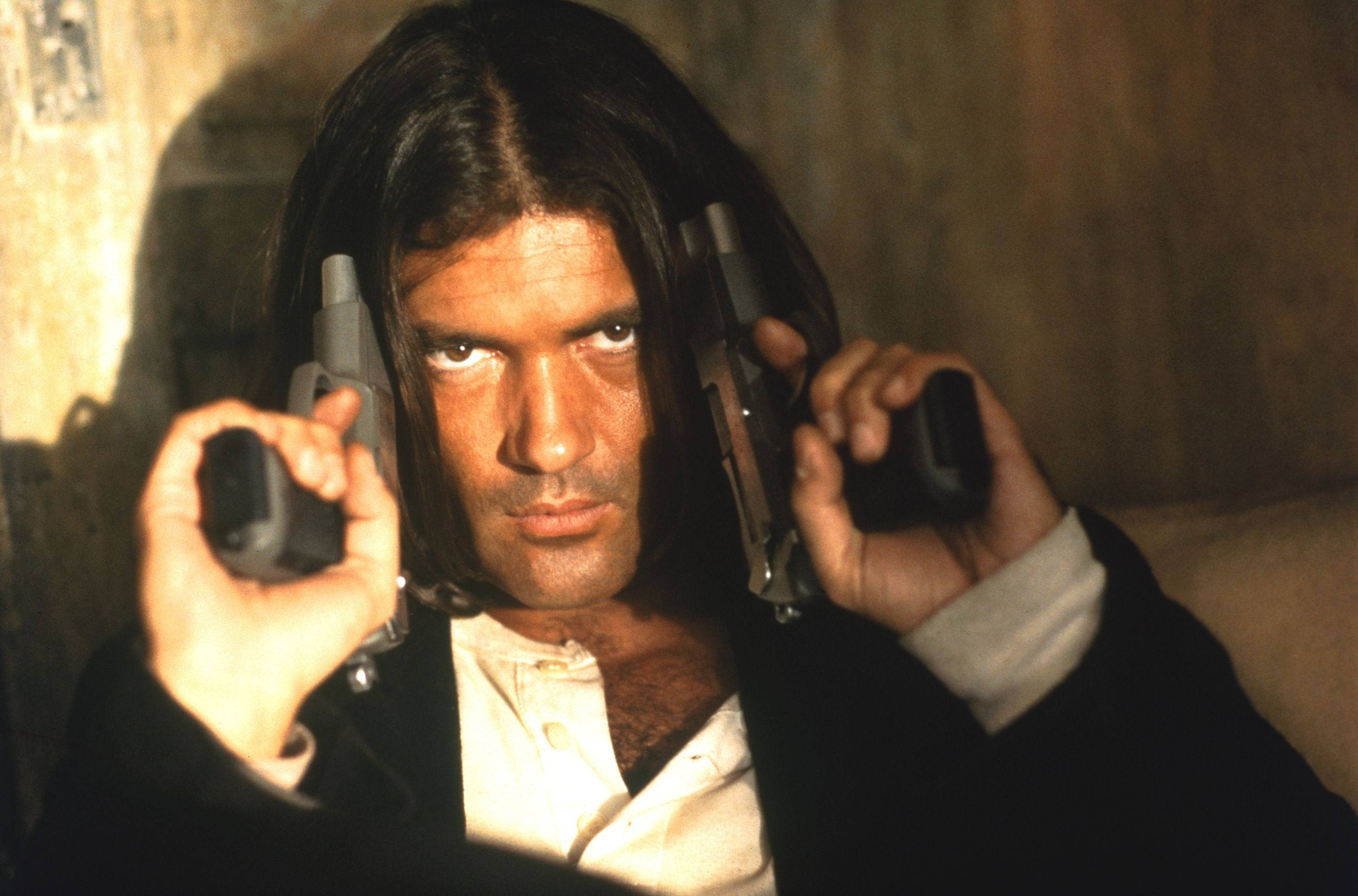 antonio banderas 桌布 壁紙 — 一名長黑髮的男子在光線昏暗、帶有暖色照明的房間中，雙手各持一把手槍指向前方。