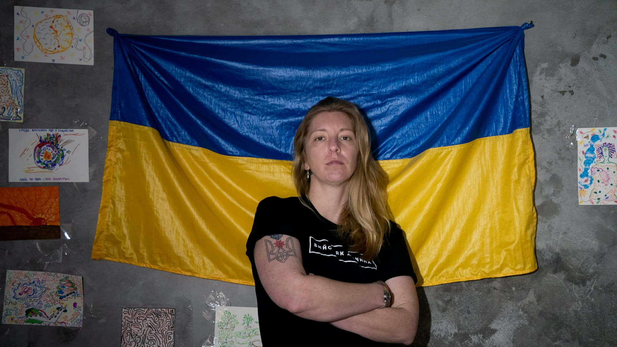 Ukraine Women 桌布 壁紙 — 一名雙臂交叉的女性莊嚴地站在一面巨大的烏克蘭國旗前，周圍是混凝土牆上的兒童藝術品。