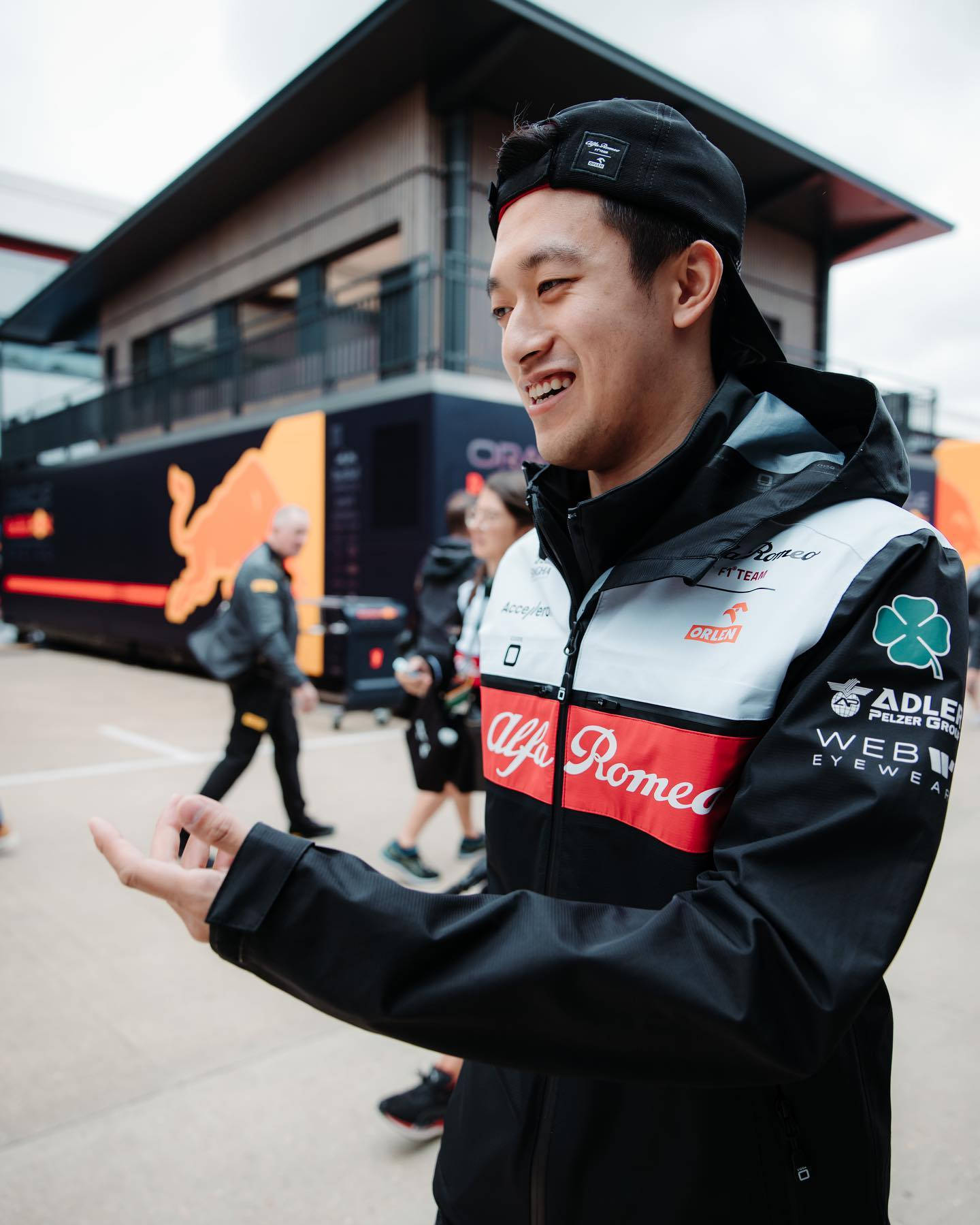 F1 桌布 壁紙 — 一名面帶微笑、身穿愛快羅密歐賽車外套的男子，在帶有車隊品牌的現代維修站車庫外愉快地打手勢。