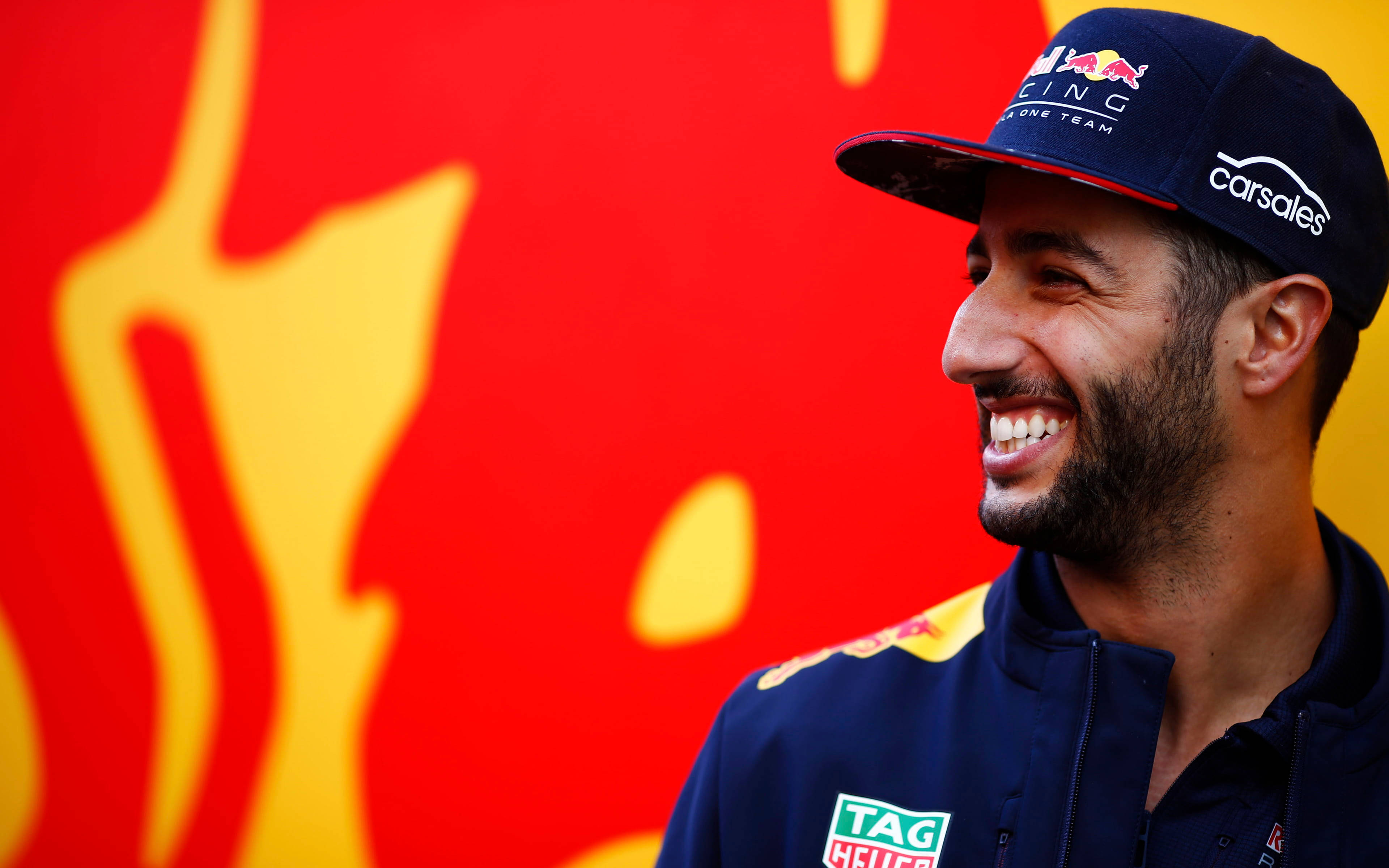 Smile 桌布 壁紙 — 一名面帶微笑的男子身穿Red Bull Racing車隊外套和帽子，站在充滿活力的紅黃抽象背景前。