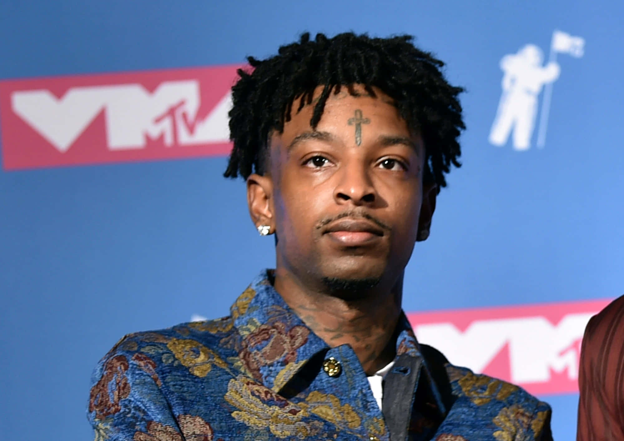 21 Savage 桌布 壁紙 — 一名額頭有十字刺青、留著辮子頭的男子，在紅毯活動中穿著一件有圖案的藍色外套。