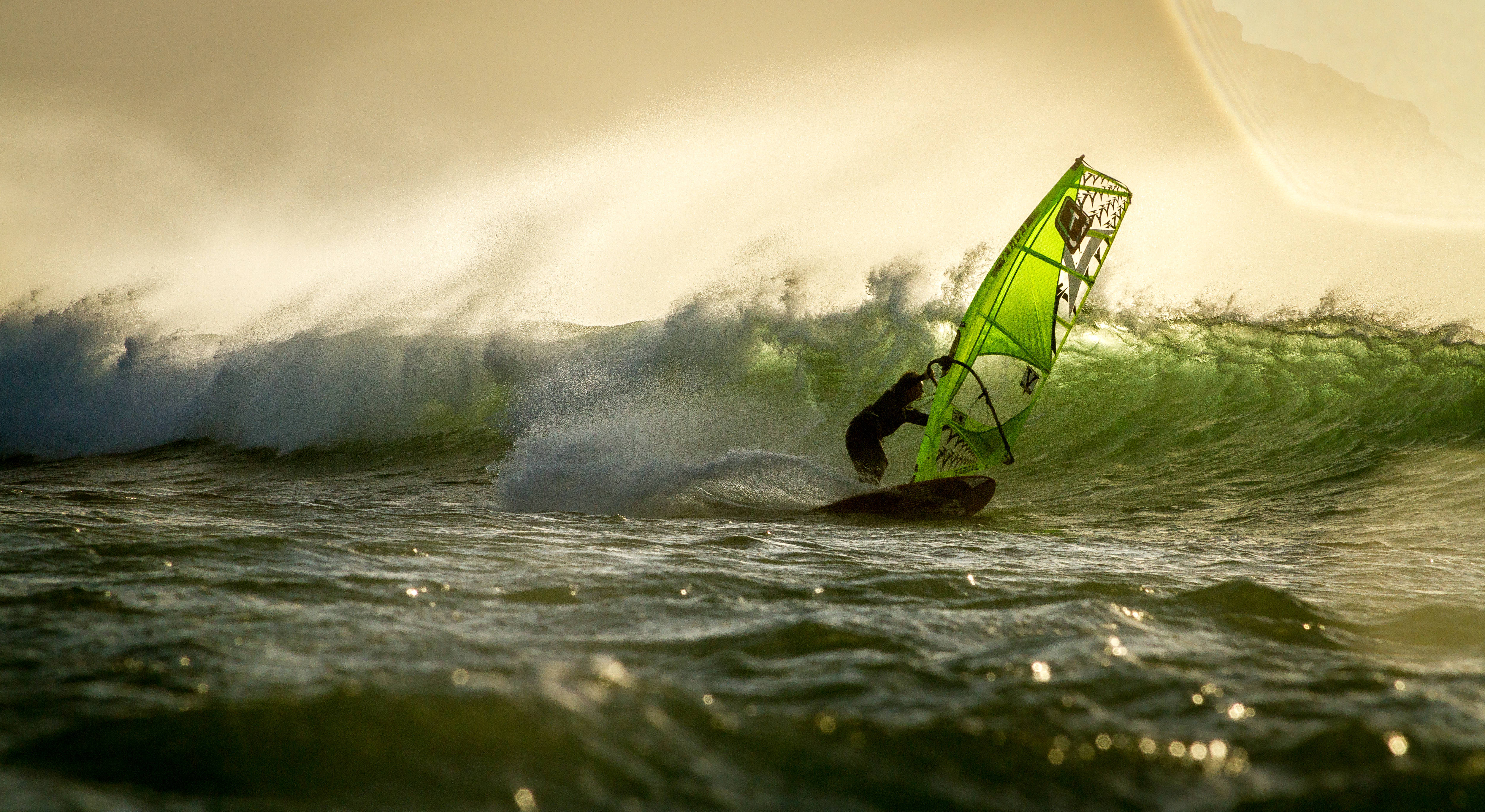 Windsurfing 桌布 壁紙 — 一名風帆衝浪者在日落時分騎乘巨大的綠色海浪，水花飛濺，金色光線從背後照亮場景。