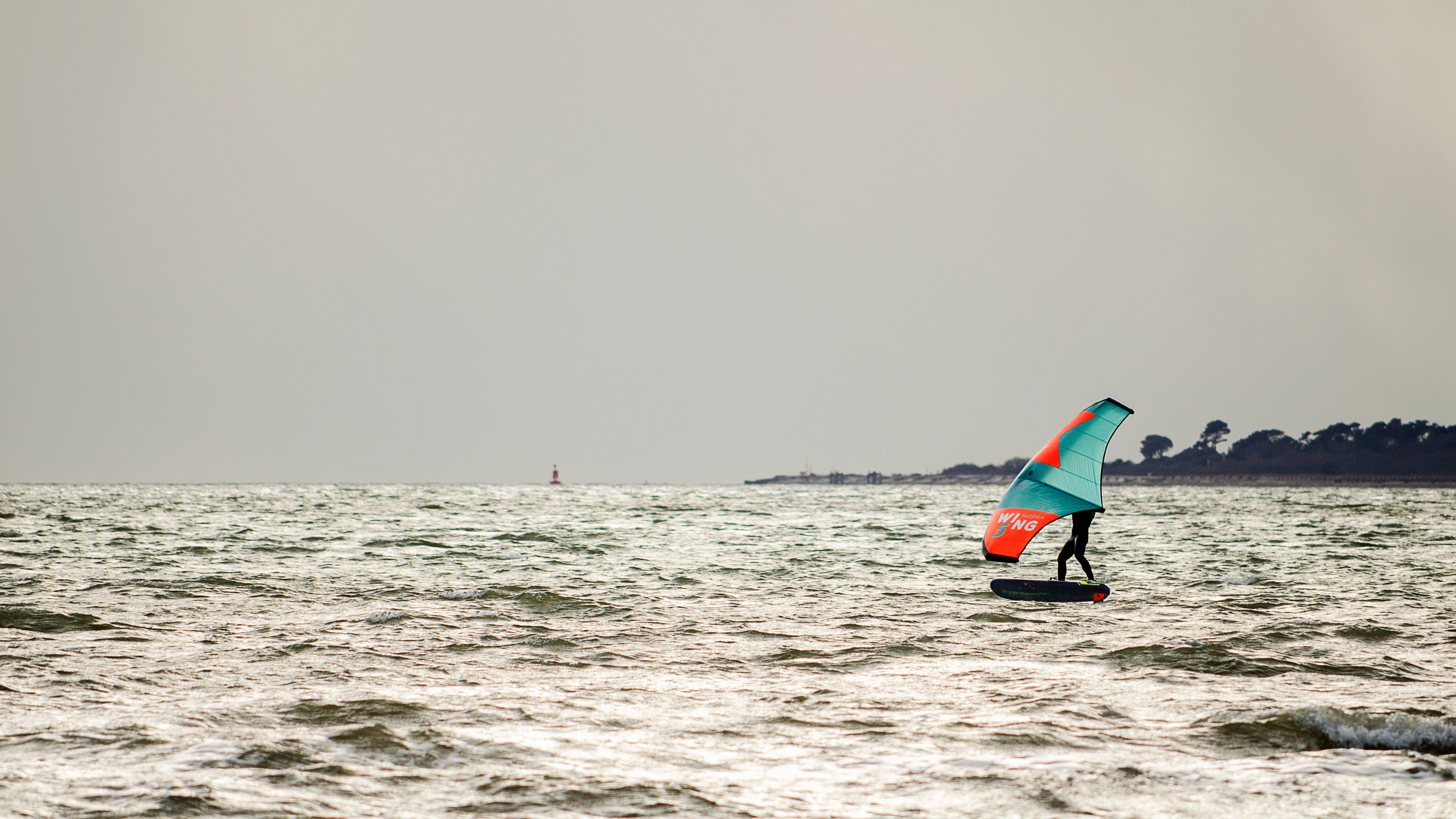 Windsurfing 桌布 壁紙 — 一名風帆選手在朦朧的天空下，於波濤洶湧的水面上滑行，遠處有海岸線和燈塔。