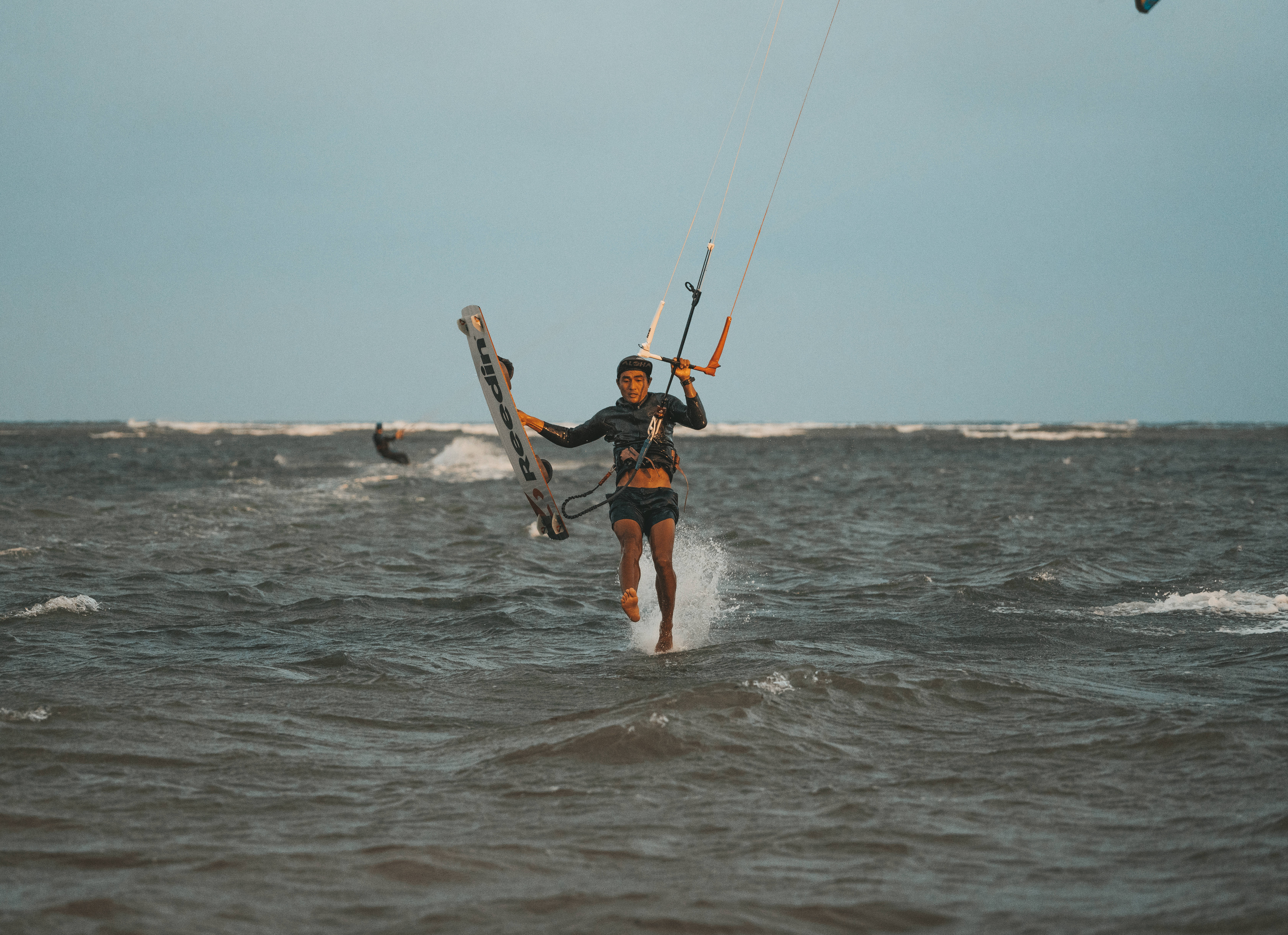 Windsurfing 桌布 壁紙 — 一名風箏衝浪手在晴朗的天空下，手持滑板和操控桿，飛越波濤洶湧的海浪。