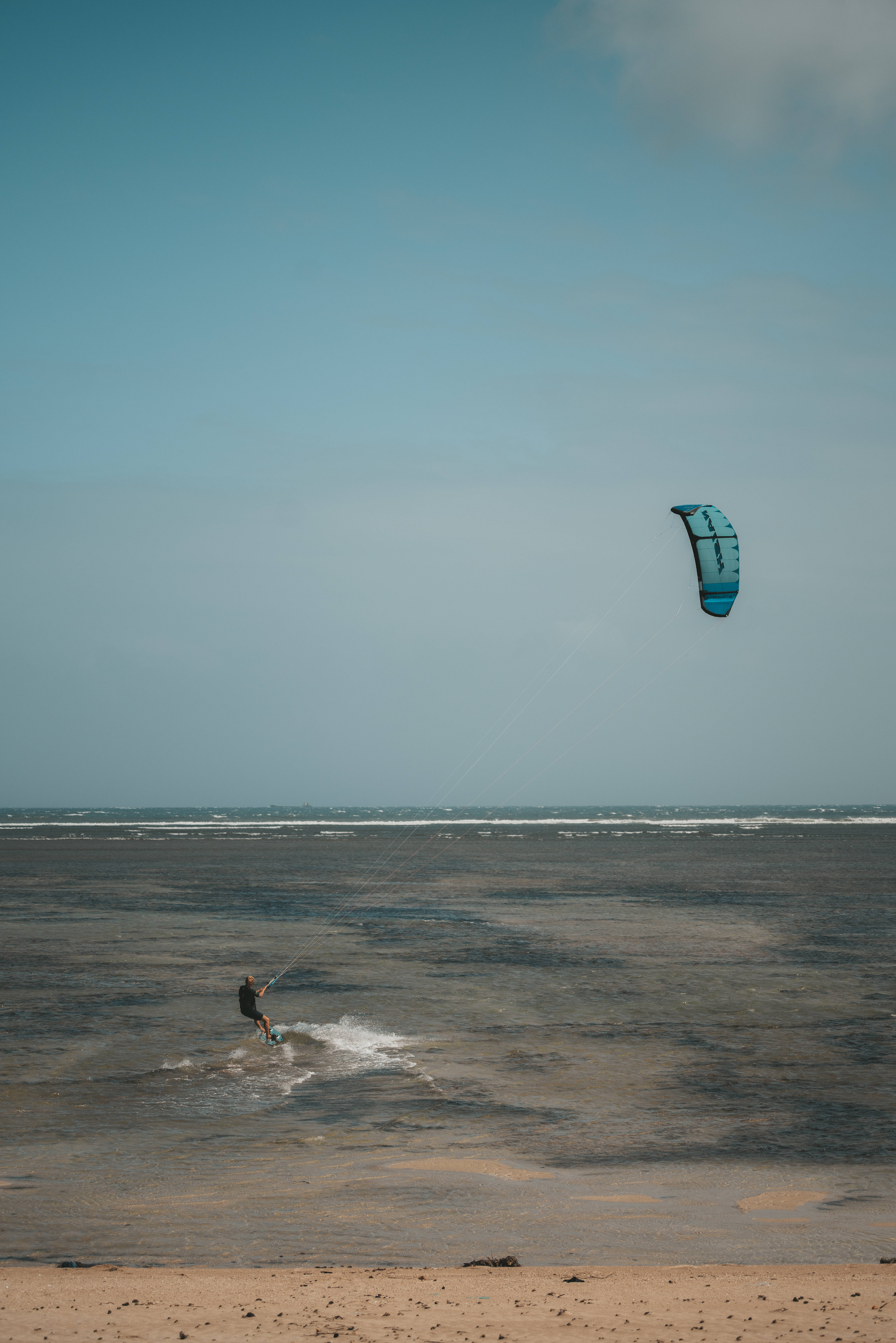 Windsurfing 桌布 壁紙 — 一名風箏衝浪者在廣闊的藍天下，於淺水沿岸水域滑行，頭頂上有一隻充滿活力的藍色風箏翱翔。