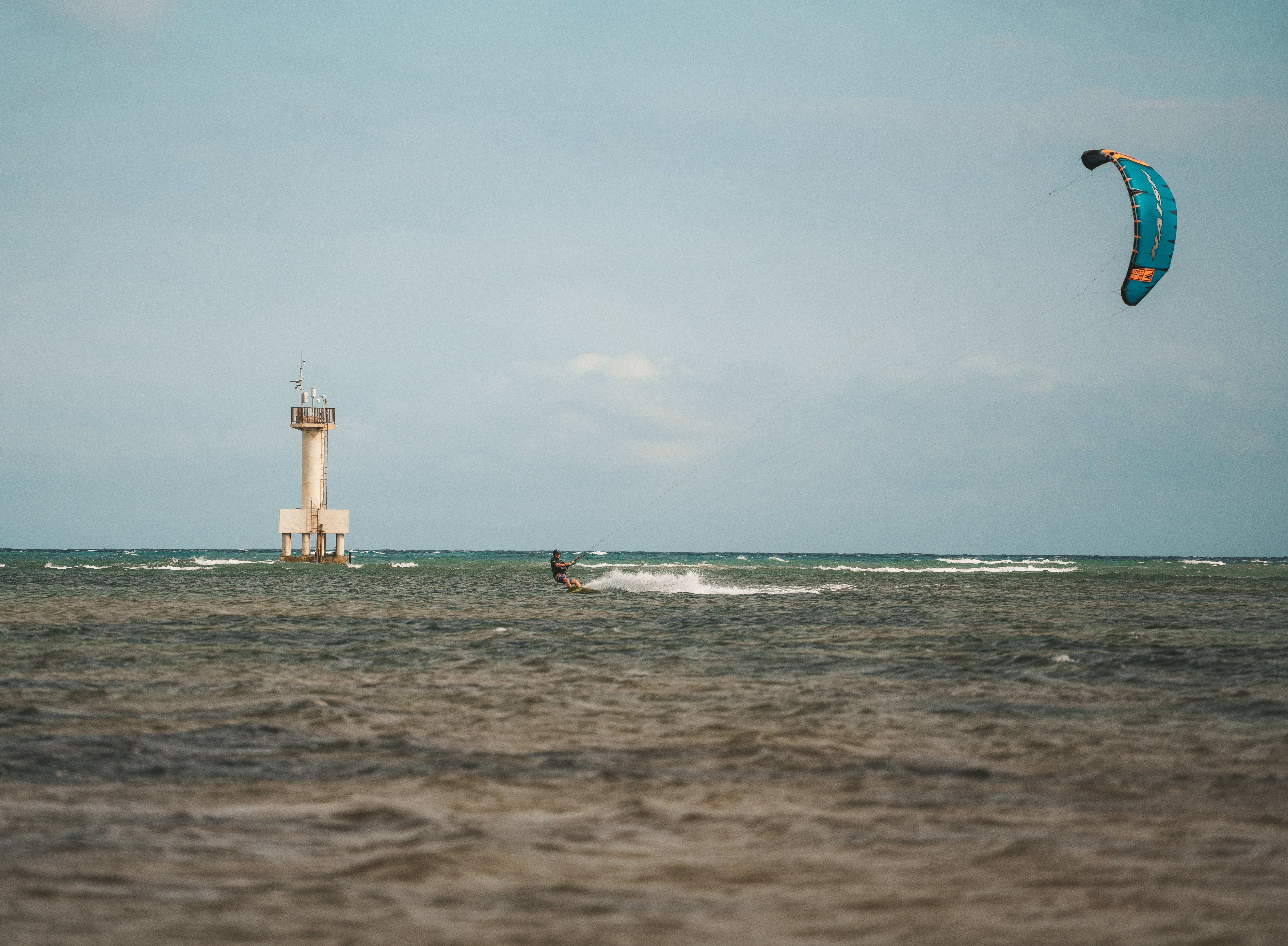 Windsurfing 桌布 壁紙 — 一名風箏衝浪者在淡藍色天空下，於波濤洶湧的海面上滑行，靠近遠處的燈塔。