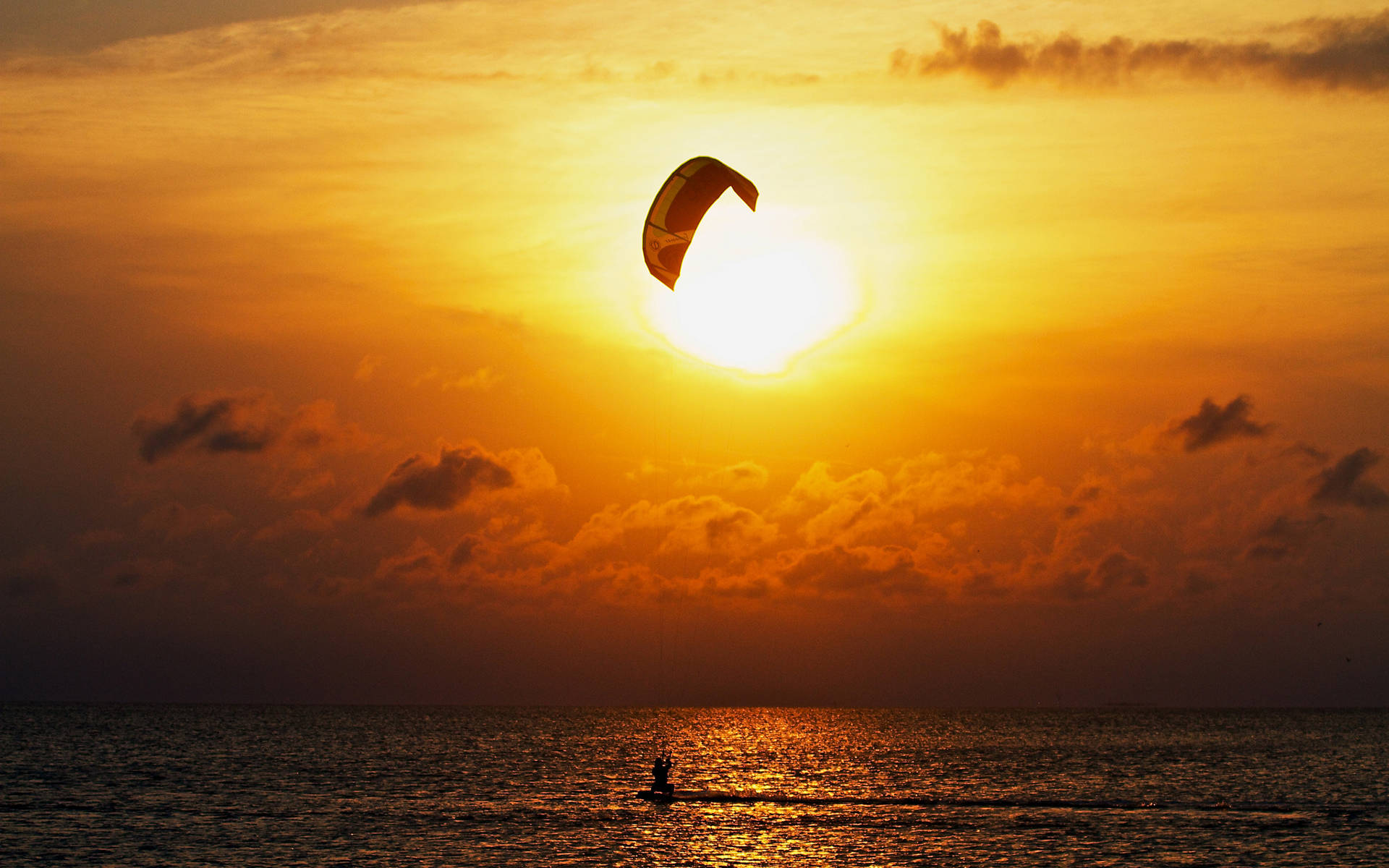 海灘 桌布 壁紙 — 一名風箏衝浪者在金色水面上滑行，夕陽在充滿活力的橙色天空後落下。