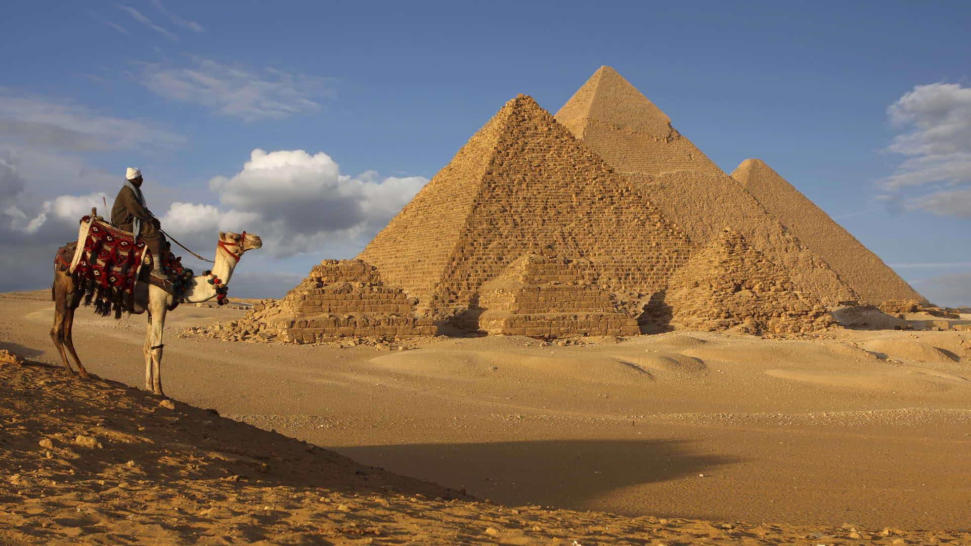 Ancient Egypt 桌布 壁紙 — 一名駱駝騎手站在雄偉的吉薩金字塔前，天空湛藍，雲朵稀疏。