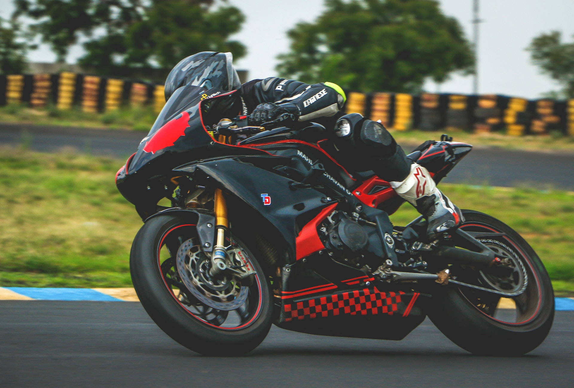 Racing 桌布 壁紙 — 一名騎士在賽道上騎著黑紅色運動機車傾斜入一個急彎，背景有模糊的綠化和黃色護欄。
