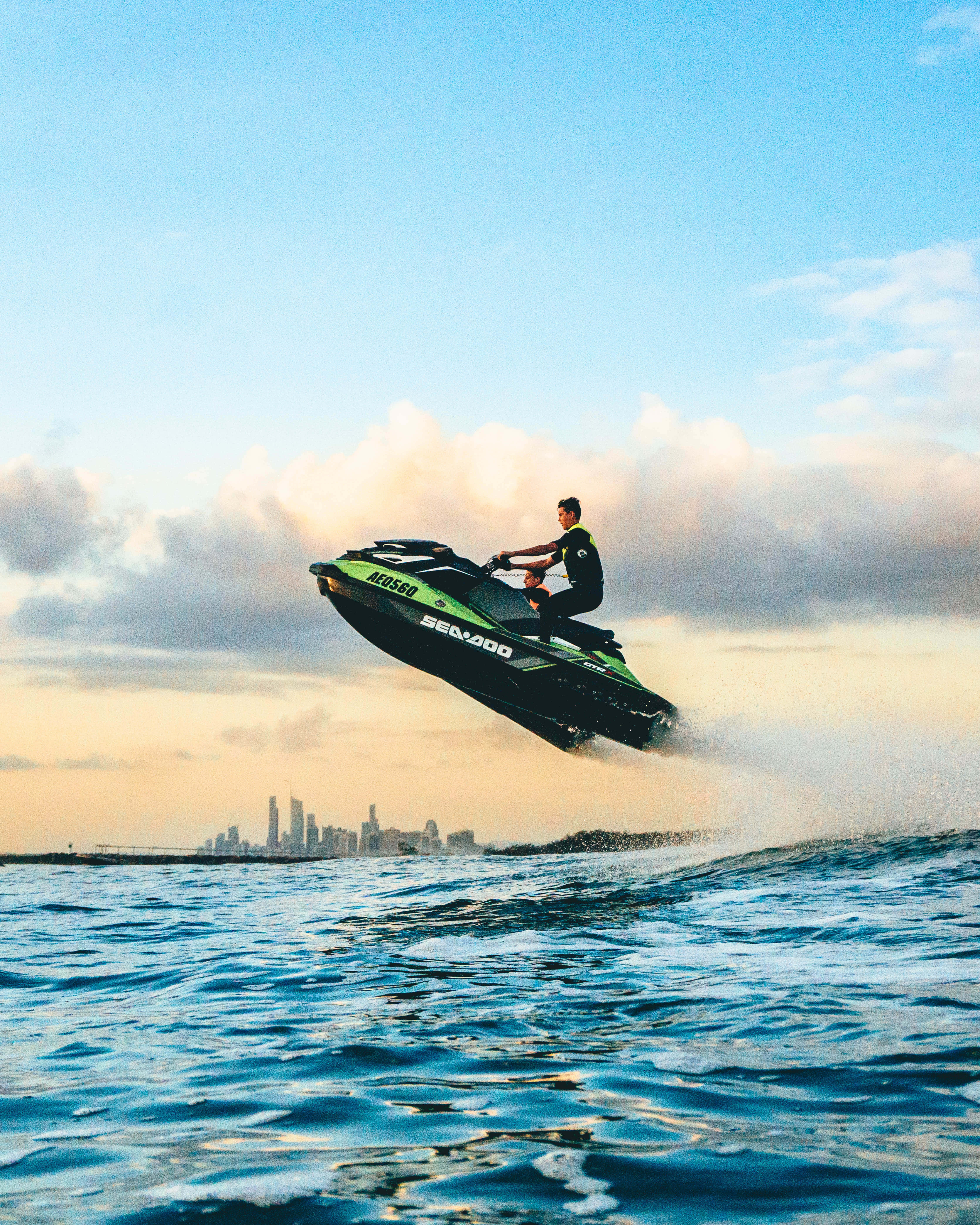 Man 桌布 壁紙 — 一名騎士駕駛綠色和黑色的 Sea-Doo 水上摩托車在海浪上奔馳，夕陽下城市天際線清晰可見。
