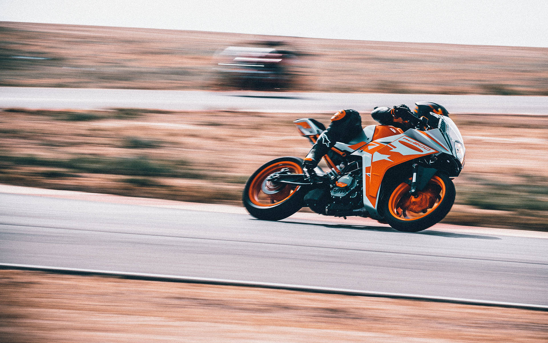 KTM RC 200 桌布 壁紙 — 一名騎士騎著橙白色運動機車，在沙漠公路上高速飛馳，並帶有動態模糊效果地傾斜入彎。