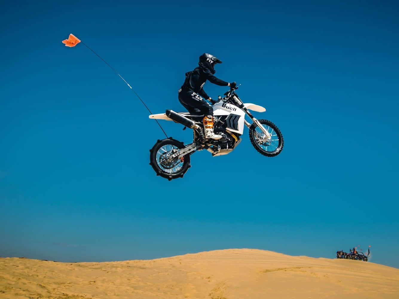 Dirtbike 桌布 壁紙 — 一名騎士騎著白色越野摩托車在金色沙丘上空飛翔，藍天白雲下，橙色旗幟在身後飄揚。