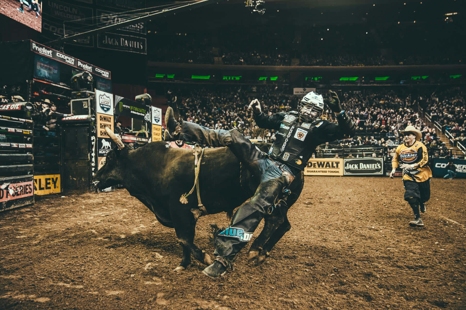 Bull Rider 桌布 壁紙 — 一名騎牛士在擠滿觀眾的牛仔競技場中，在明亮的體育場燈光下緊緊抓住一頭狂跳的公牛。