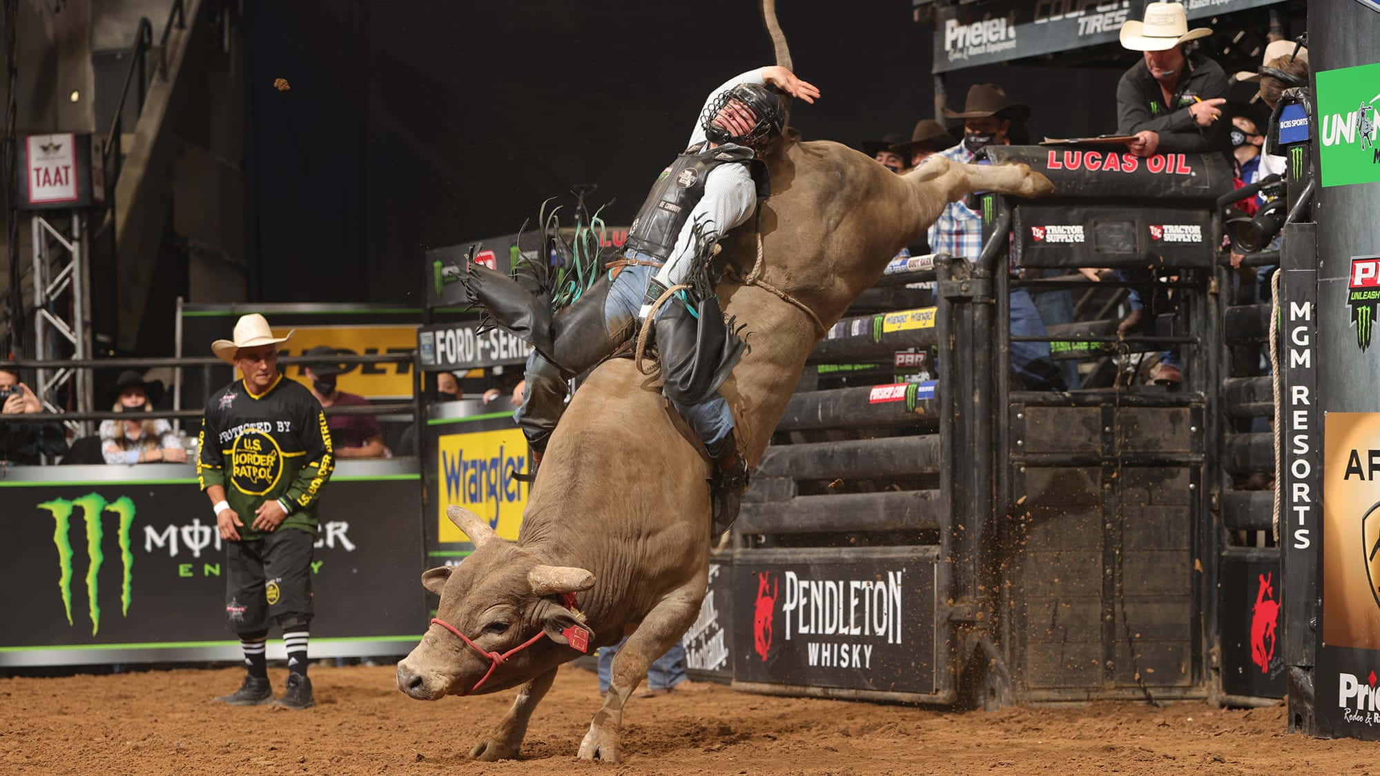 Bull Riding 桌布 壁紙 — 一名騎牛士在牛仔競技場中緊抓著一頭高高躍起的蠻牛，場內有觀眾和印有品牌的圍欄。