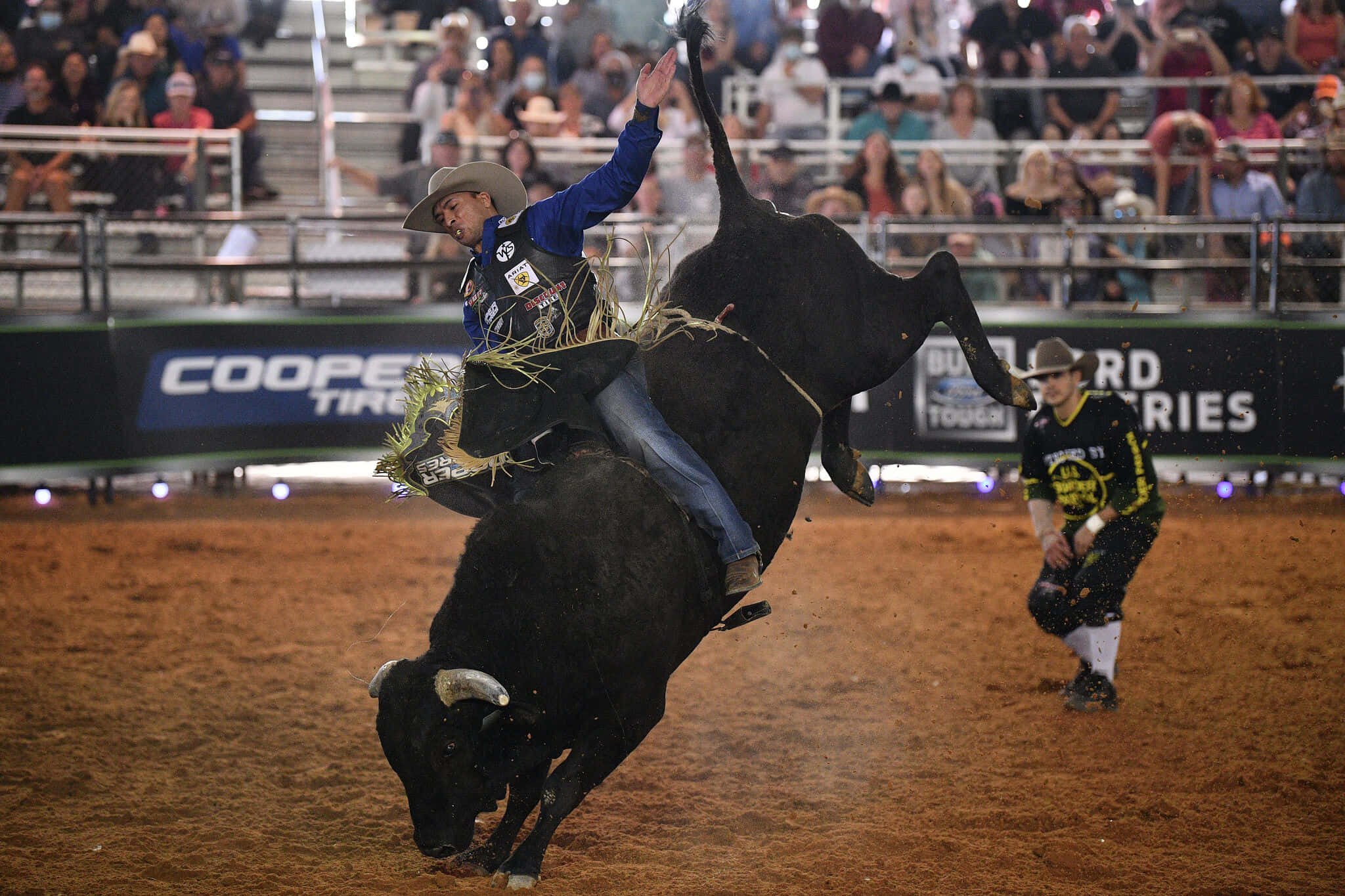 Bull Riding 桌布 壁紙 — 一名騎牛士在牛仔競技場緊緊抓住一頭正在狂跳的黑牛，旁邊有一位牛仔在觀看。