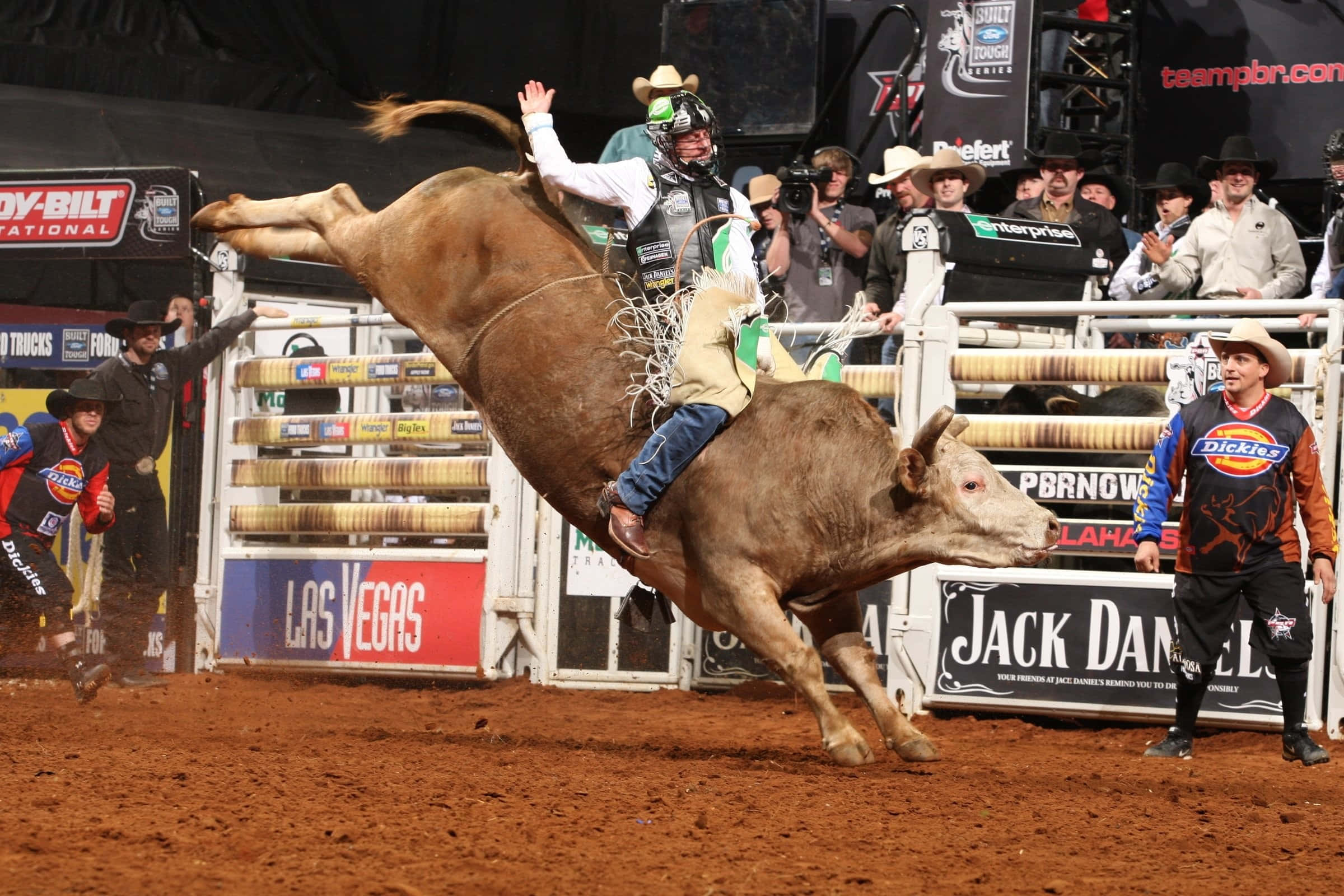 Bull Rider 桌布 壁紙 — 一名騎牛選手緊抓著一頭在拉斯維加斯牛仔競技場中猛烈甩動的棕色公牛，觀眾在欄杆後觀看。