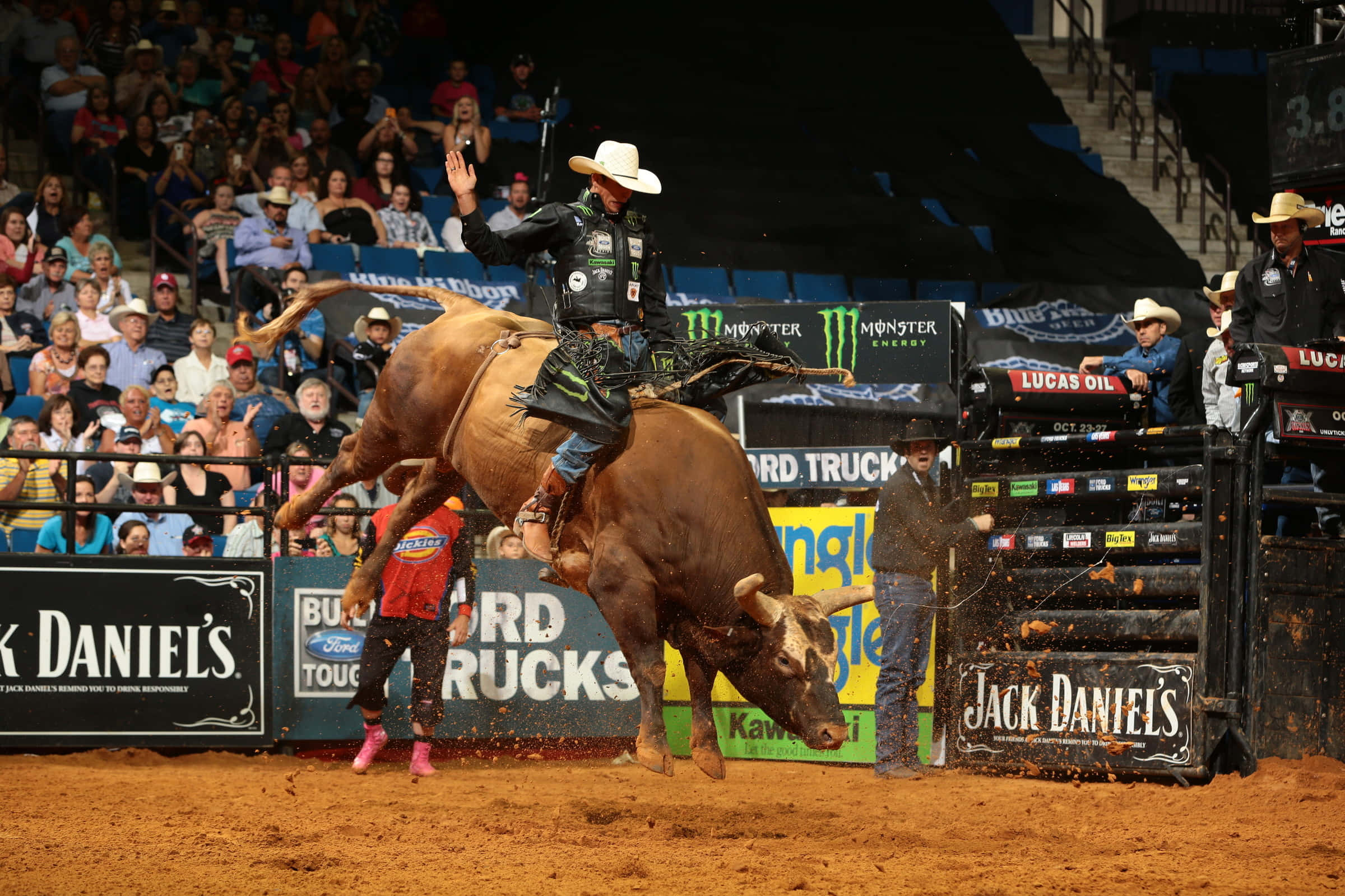 Bull Rider 桌布 壁紙 — 一名騎牛選手緊抓著一頭在牛仔競技場中狂跳的棕色公牛，泥土飛揚，觀眾從看台上觀看。