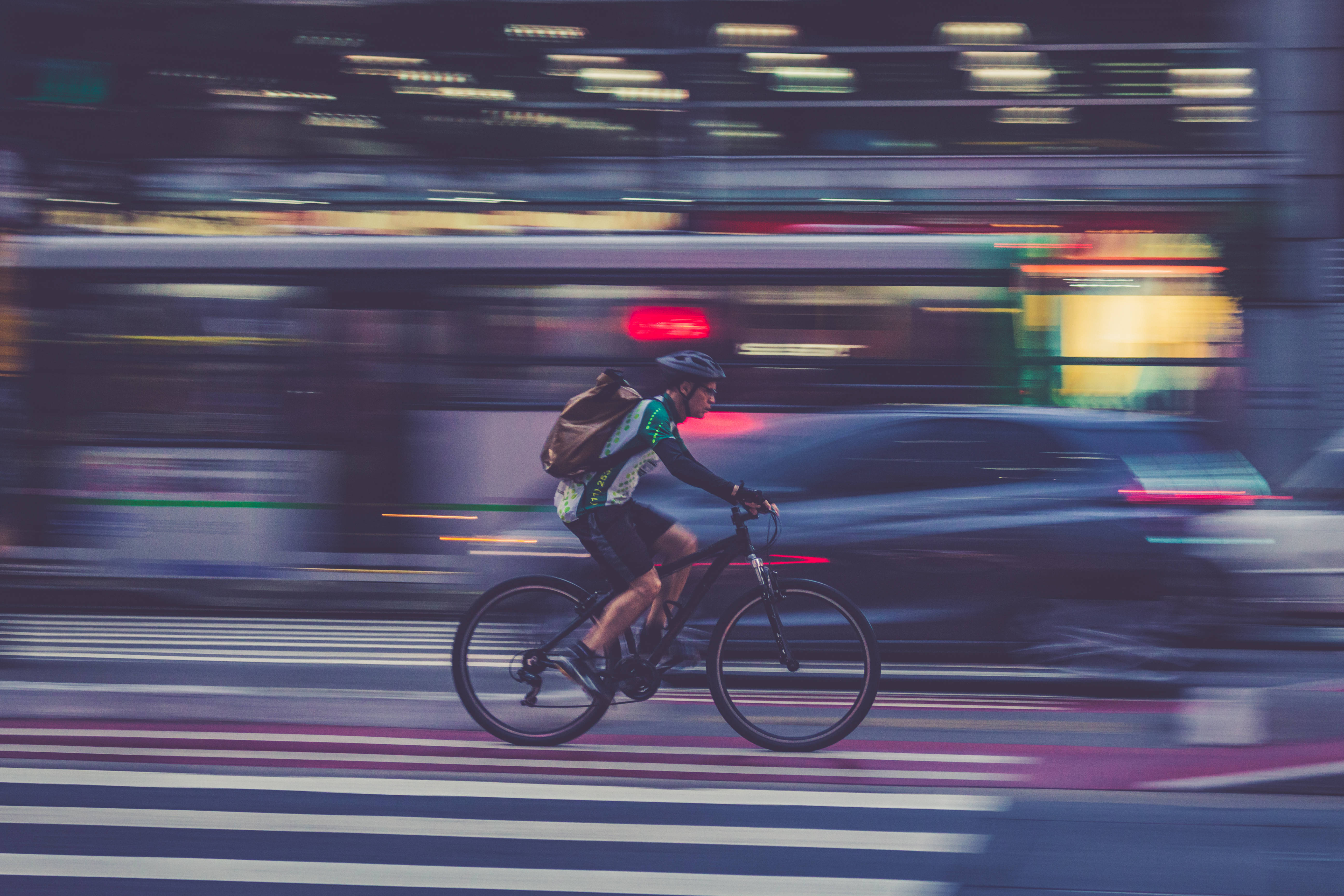 腳踏車 桌布 壁紙 — 一名騎自行車的人在夜晚穿過模糊的城市街道，伴隨著過往車輛的燈光條紋。