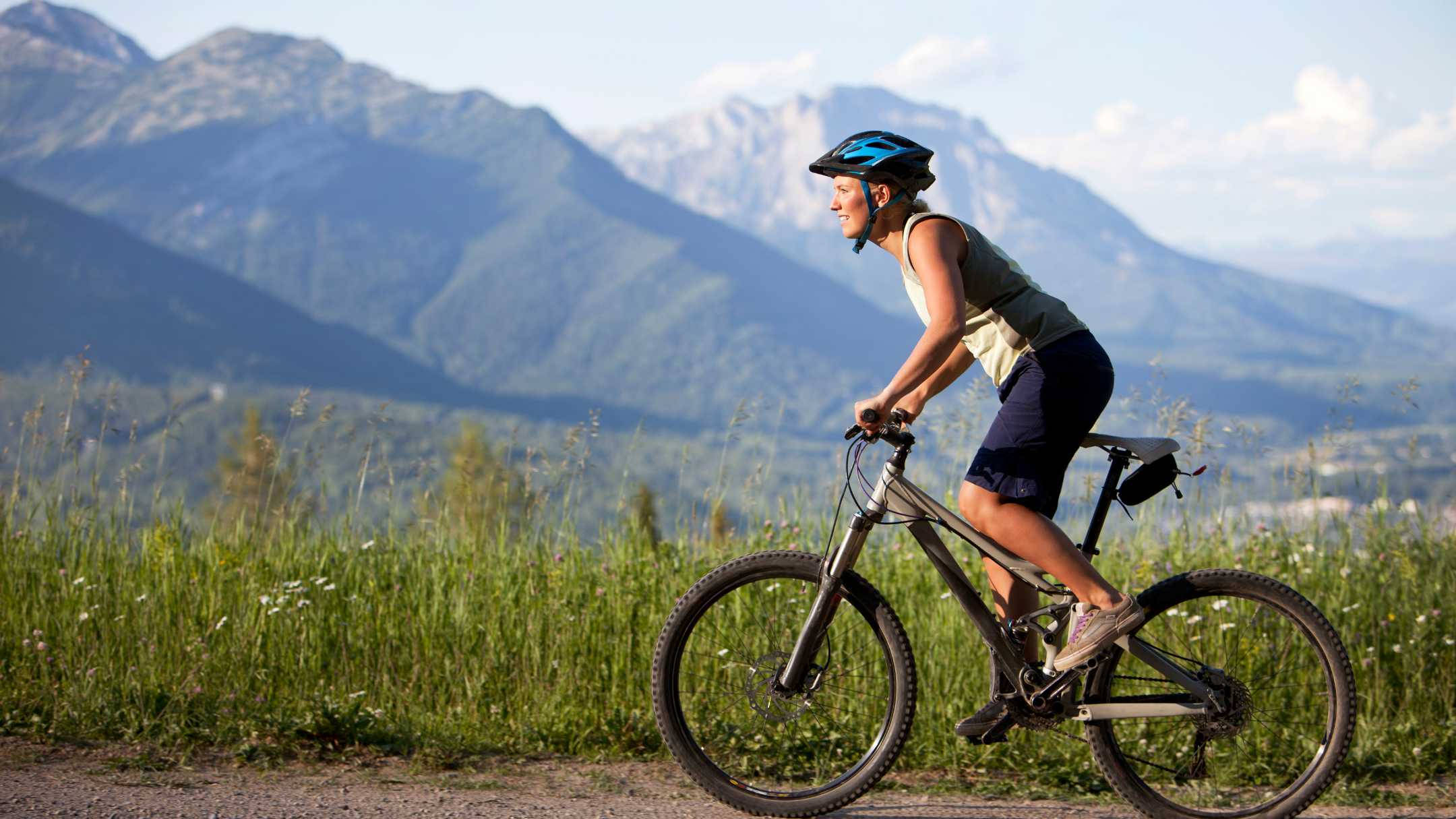 Mountain Bike 桌布 壁紙 — 一名騎自行車的人在碎石小徑上踩著登山車，背後是雄偉的雪山，聳立在茂密的綠色草地上，天空明亮。