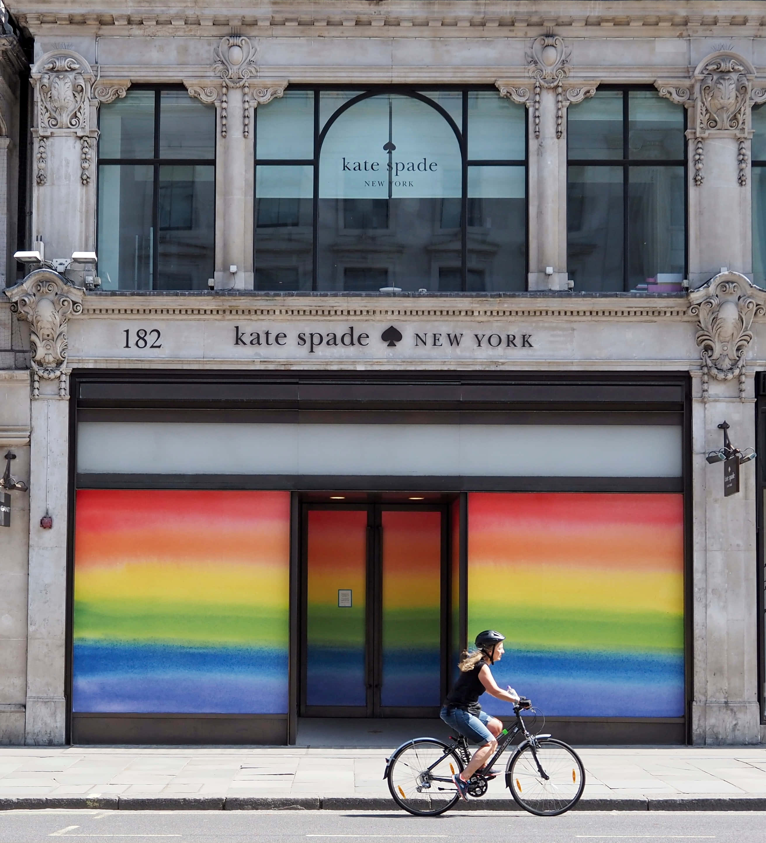 Kate Spade 桌布 壁紙 — 一名騎自行車的人經過一間Kate Spade New York商店，該商店在華麗的石造建築下設有充滿活力的彩虹色櫥窗。