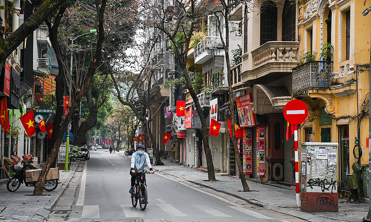 Hanoi 桌布 壁紙 — 一名騎自行車者在越南一條綠樹成蔭的城市街道上騎行，兩旁是殖民風格建築，紅旗在微風中飄揚。