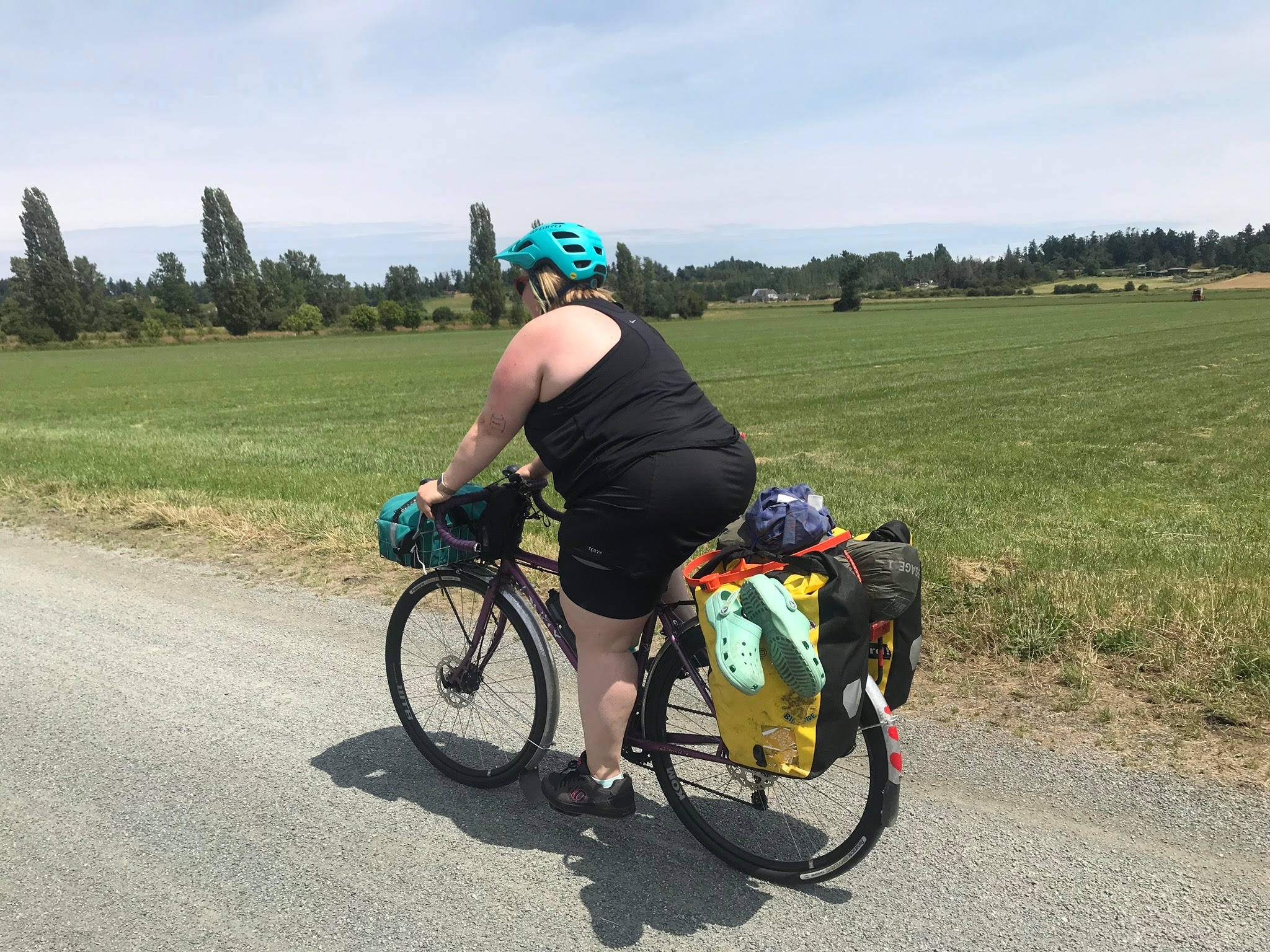 Mountain Bike 桌布 壁紙 — 一名騎自行車者戴著青綠色頭盔，踩著載滿行李的旅行自行車，沿著碎石路穿過廣闊的綠色田野，在明亮的藍天下。