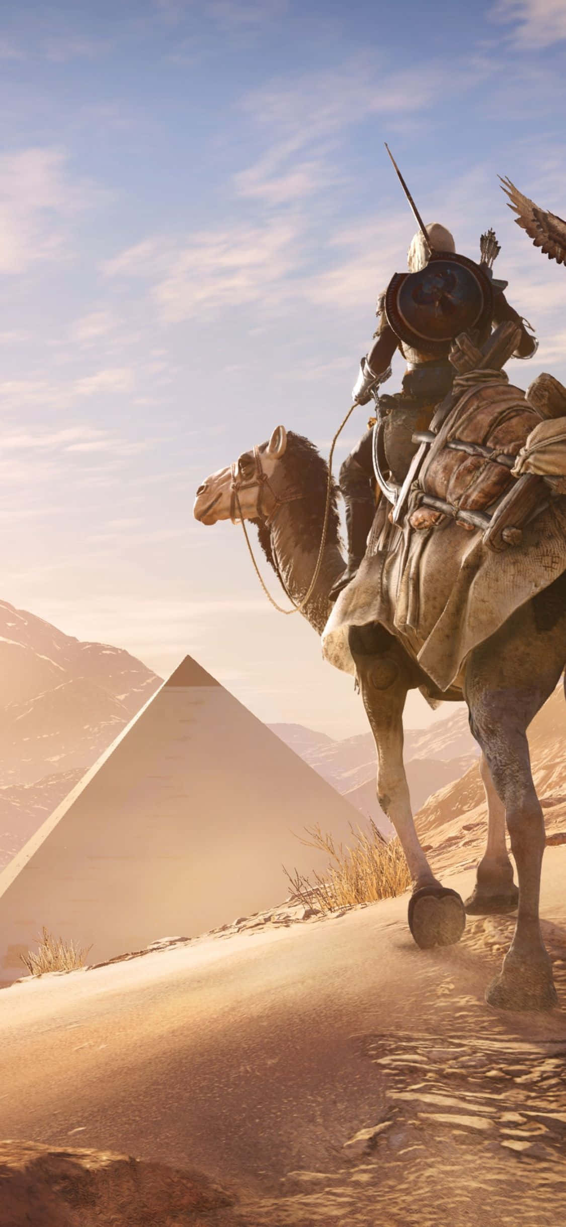 iPhone X Assassin's Creed Origins 桌布 壁紙 — 一名騎著駱駝的騎士站在沙丘頂上，在藍天下金色的沙漠薄霧中有一座金字塔。