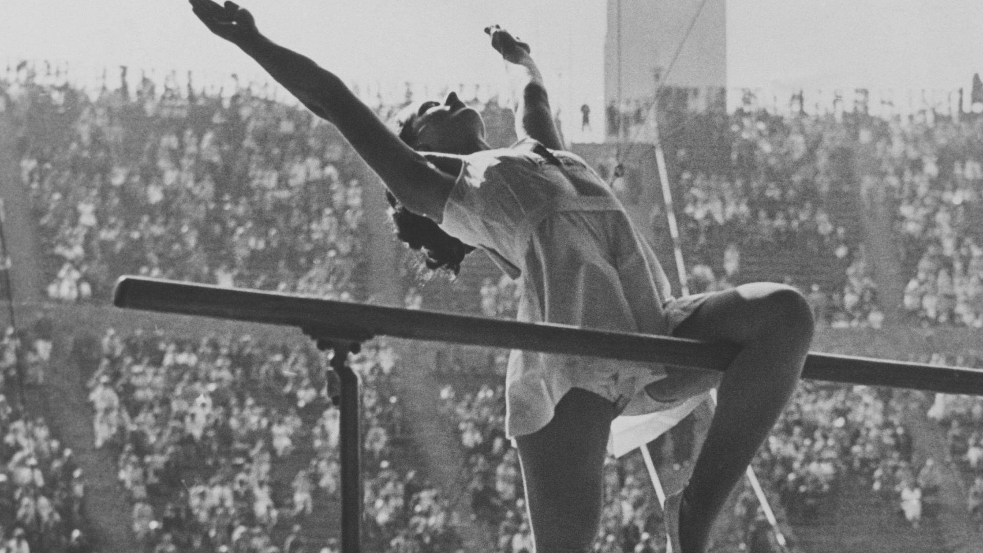 Olympics 桌布 壁紙 — 一名體操運動員在陽光普照、坐滿觀眾的體育館內，優雅地在平衡木上拱起身體。