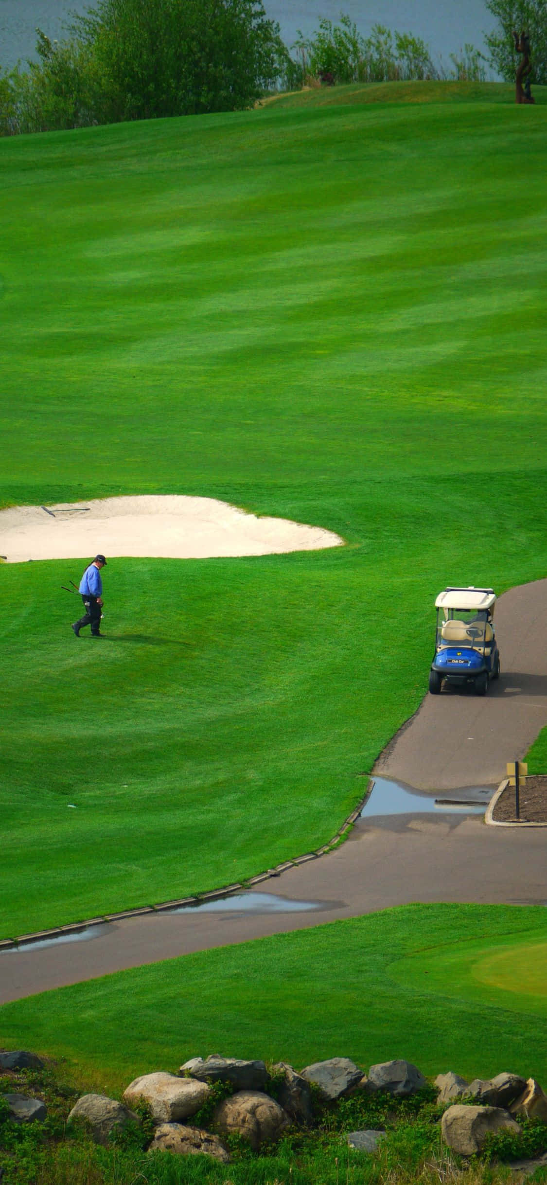 iPhone X Golf Course 桌布 壁紙 — 一名高爾夫球手在明亮的日光下，沿著充滿活力的綠色球道行走，旁邊有一輛藍色高爾夫球車和沙坑。