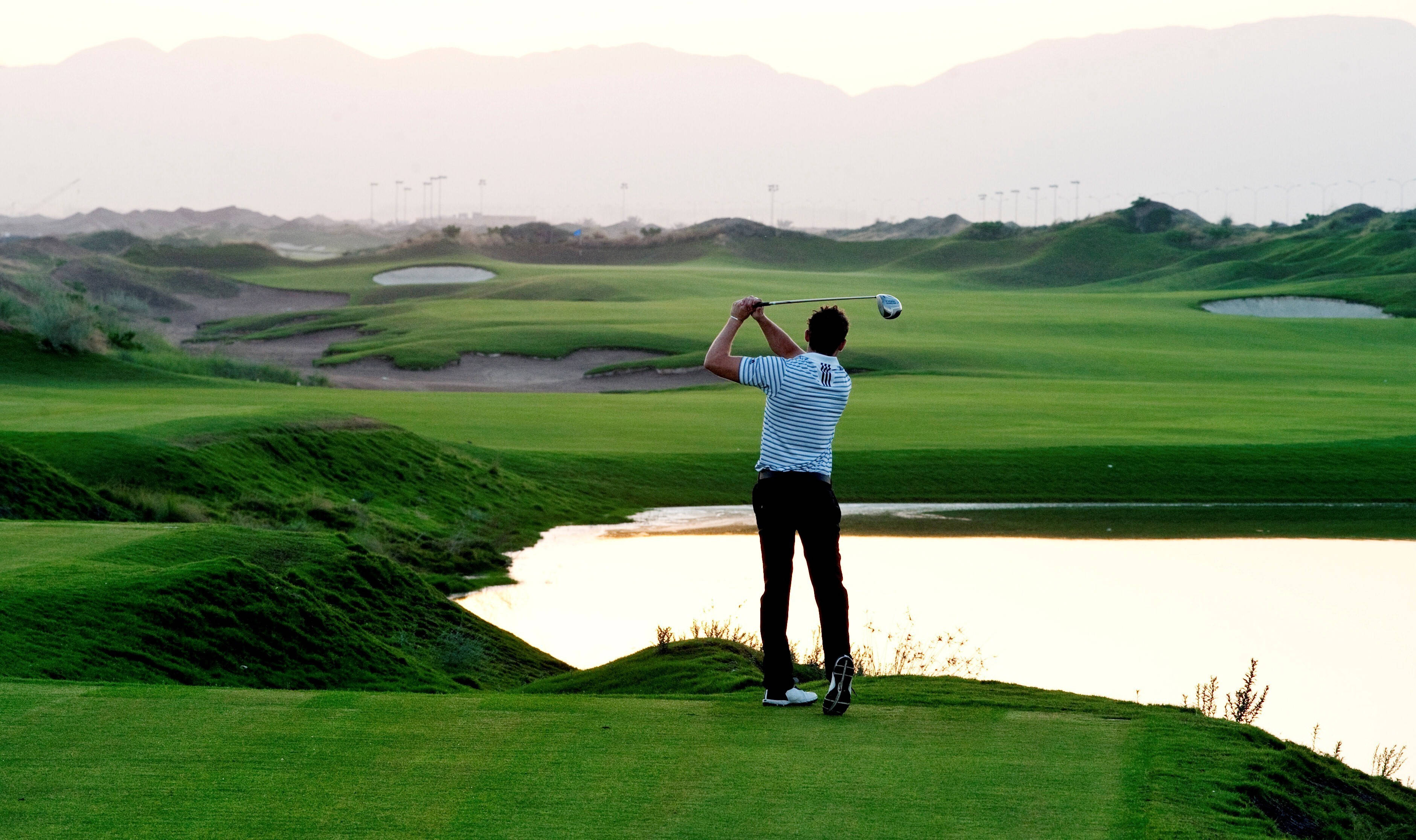 Golfing 桌布 壁紙 — 一名高爾夫球手在柔和的金色光線下，於翠綠的球場上揮桿，場地有水域障礙和遠山。