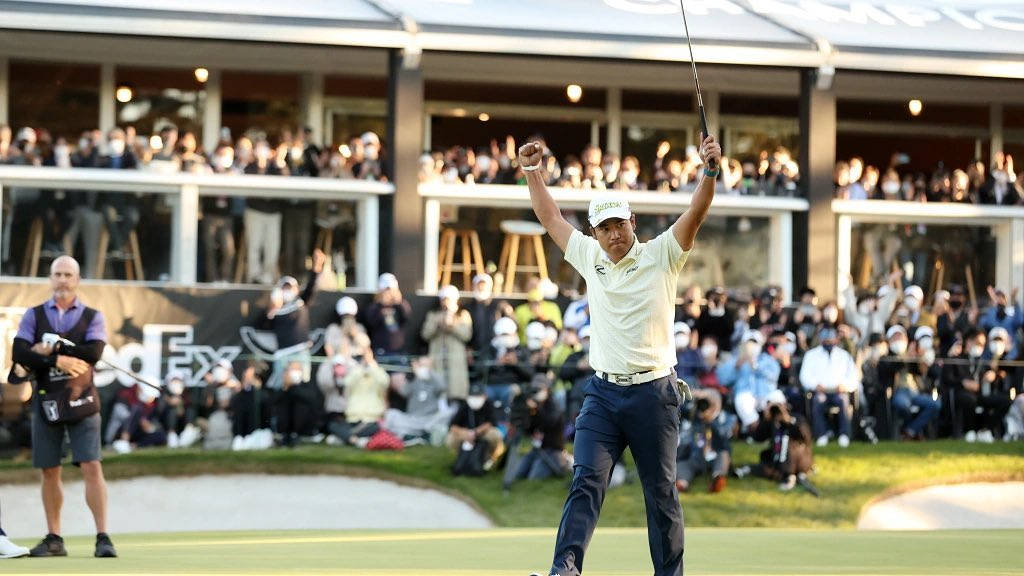 Champion 桌布 壁紙 — 一名高爾夫球手在陽光明媚的果嶺上勝利地高舉雙臂和球桿，而歡呼的觀眾從陽台和球道觀看。