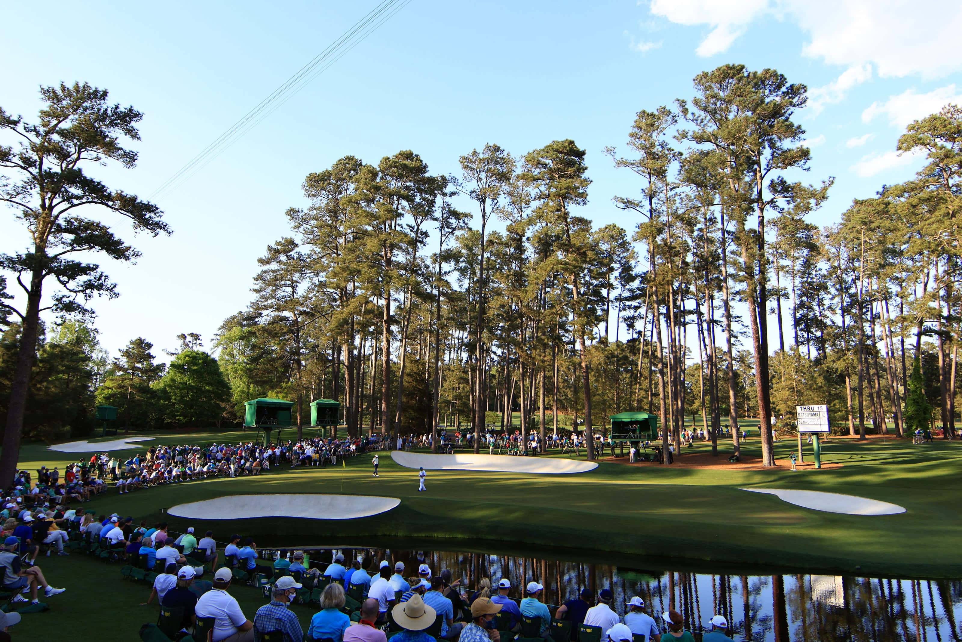 Augusta National iPhone 桌布 壁紙 — 一名高爾夫球手在陽光明媚的球道上開球，周圍是高聳的松樹，觀眾沿著水障礙區就座。