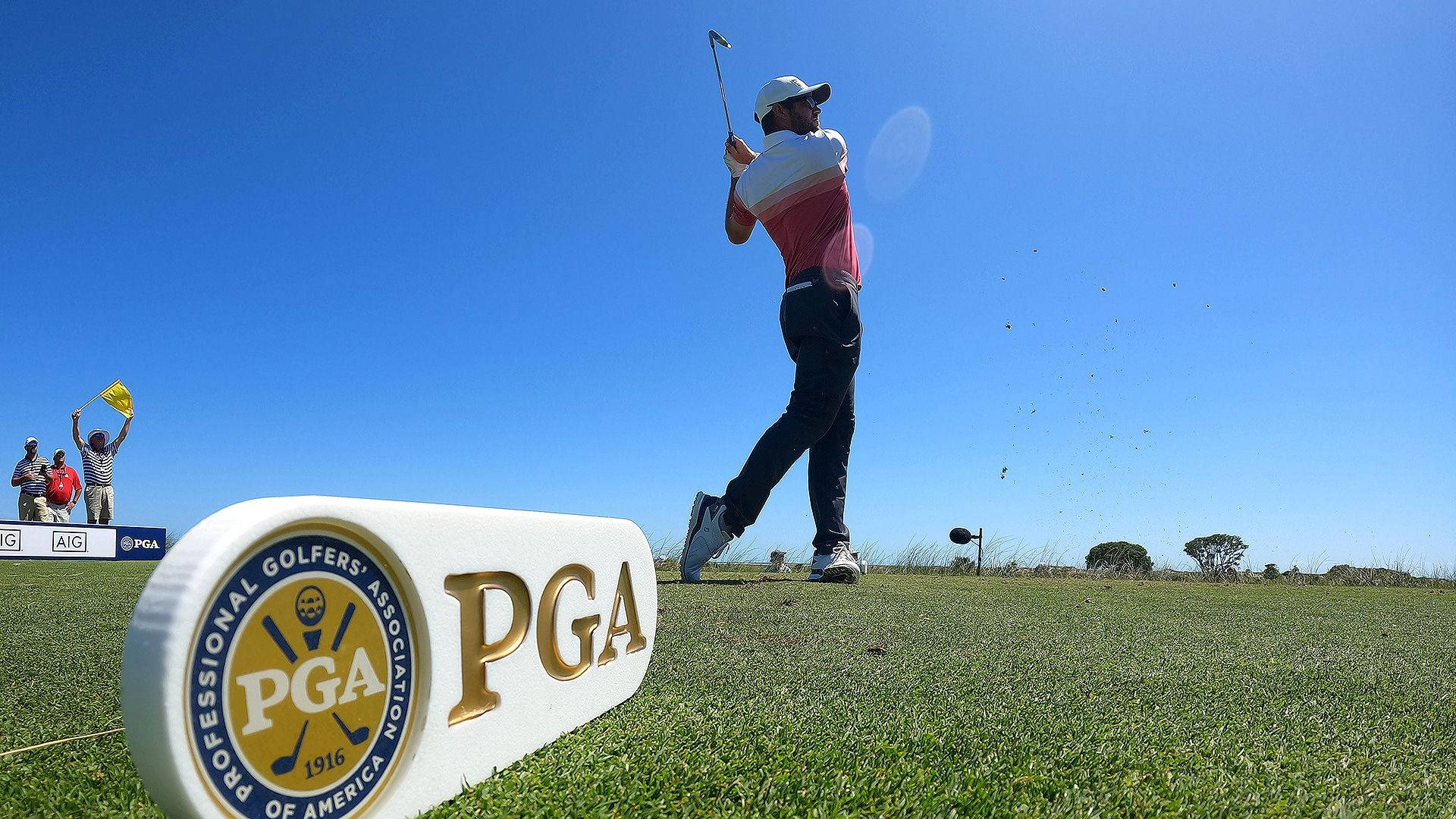 Pga 桌布 壁紙 — 一名高爾夫球手在陽光明媚的球道上強力揮桿，前景有PGA標誌標記，背景有觀眾歡呼。