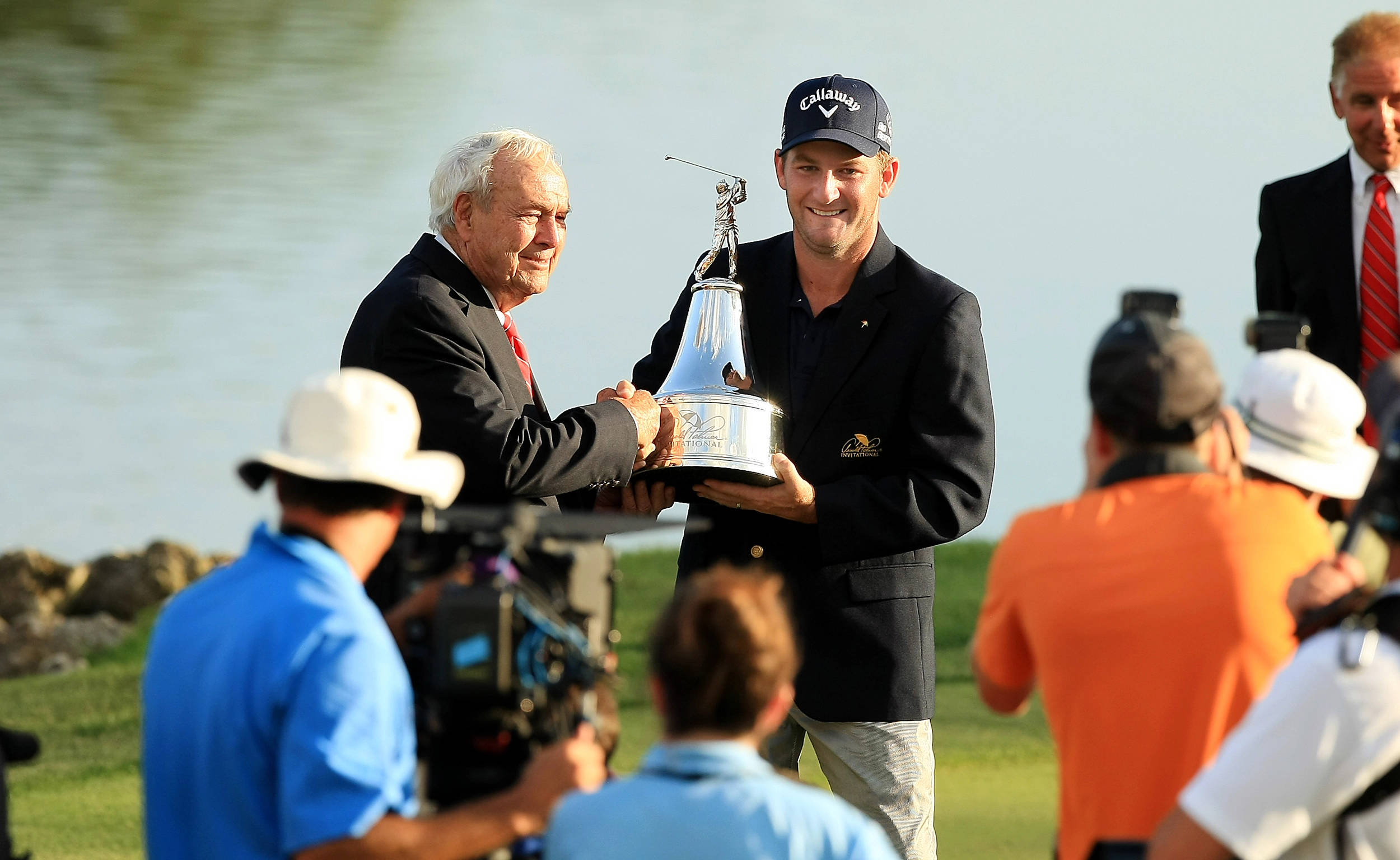 Champion 桌布 壁紙 — 一名高爾夫球手在陽光明媚的高爾夫球場上，從一位年長男子手中接過閃亮的獎盃，周圍有攝影師圍繞，他面帶微笑。
