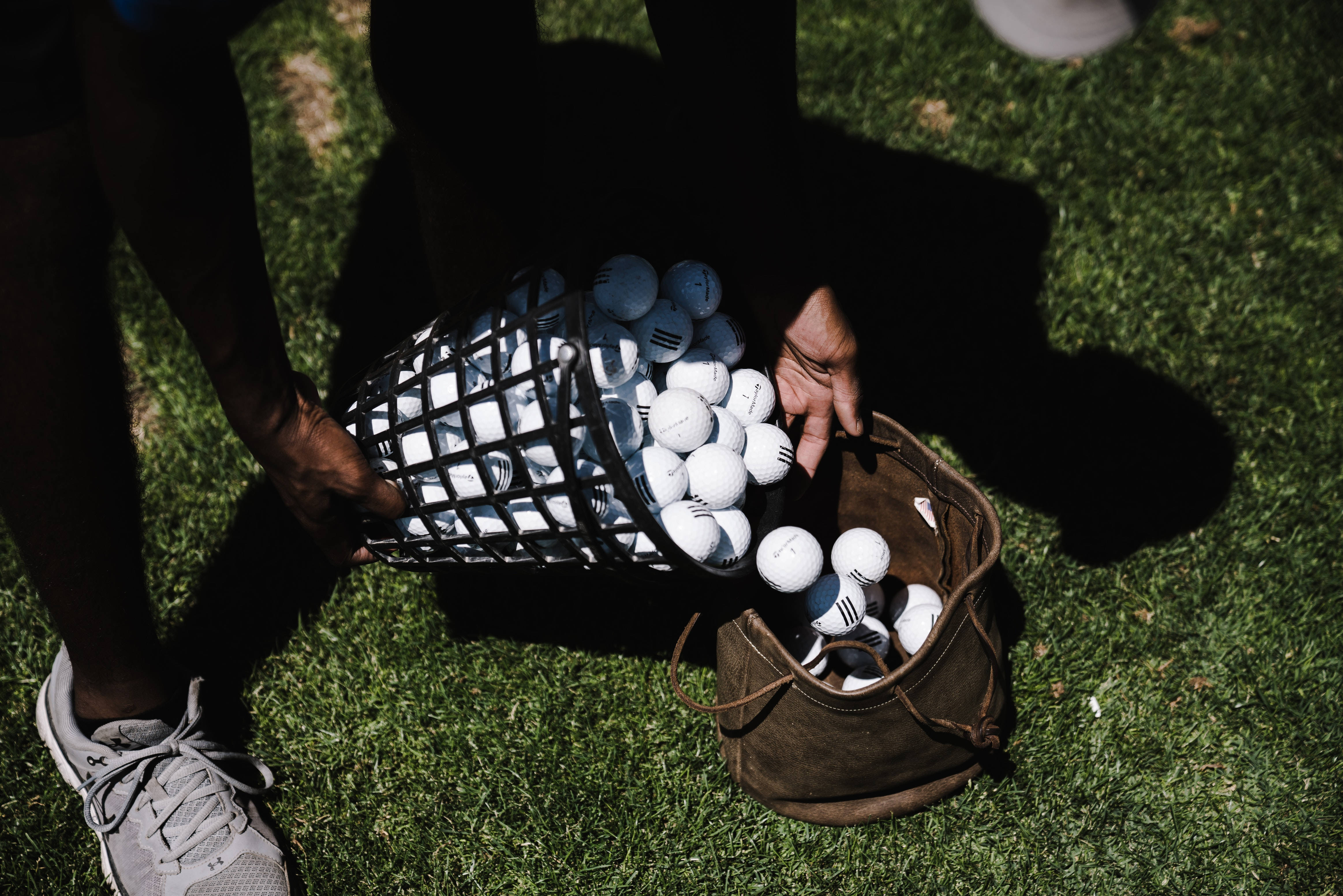 4K Golf 桌布 壁紙 — 一名高爾夫球手將白色高爾夫球從黑色籃子倒入陽光照射的綠色草地上的棕色皮革袋中。