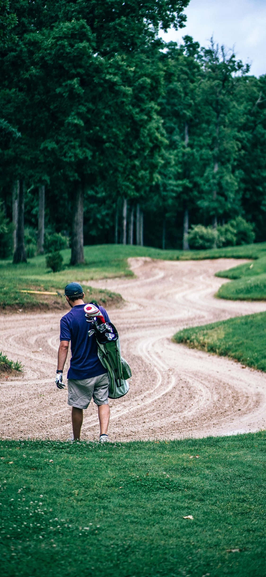 iPhone X Golf Course 桌布 壁紙 — 一名高爾夫球手沿著蜿蜒的泥土小徑穿過高爾夫球場上茂密的綠樹走開。