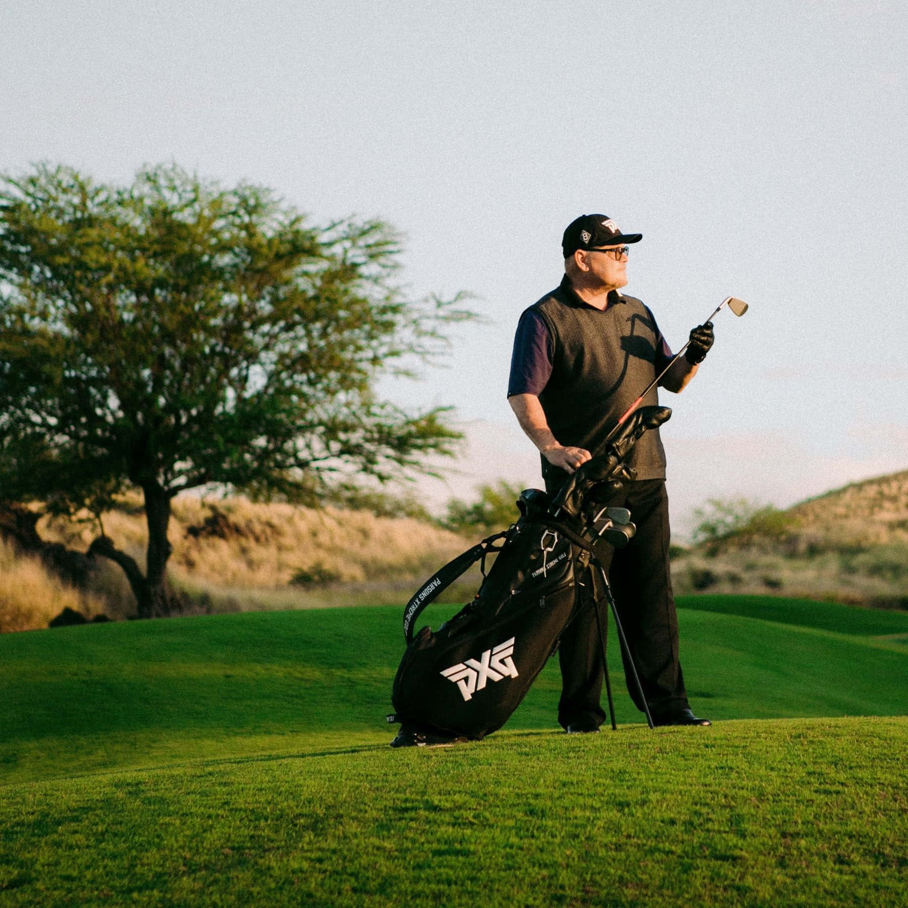 Golf Club 桌布 壁紙 — 一名高爾夫球手站在陽光明媚的綠色球場上，手持球桿，身旁放著一個PXG球袋，天空晴朗。