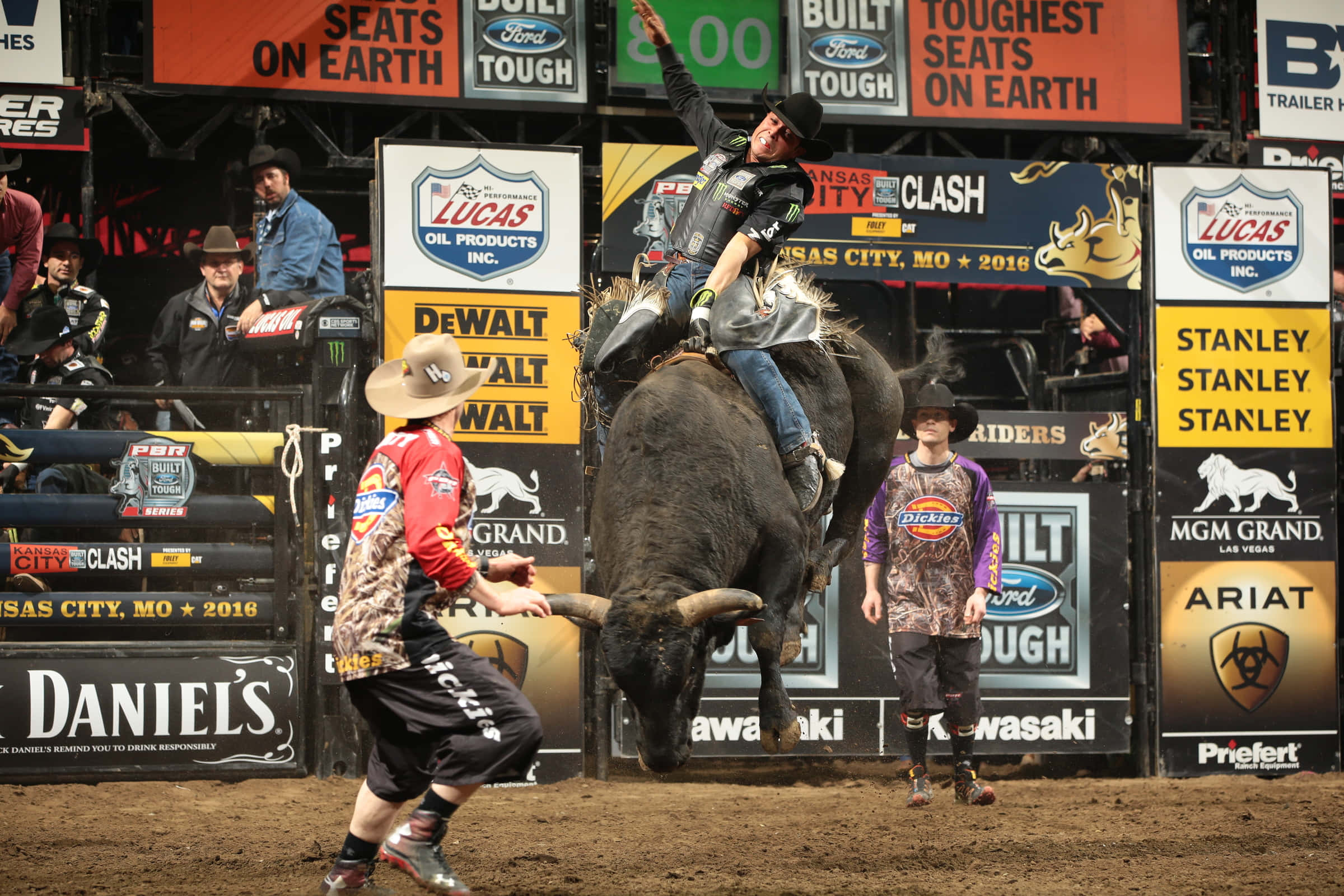Bull Riding 桌布 壁紙 — 一名鬥牛騎手緊貼著一頭猛烈踢蹬的黑牛，而牛仔小丑在明亮的燈光下於擠滿觀眾的競技場中分散其注意力。
