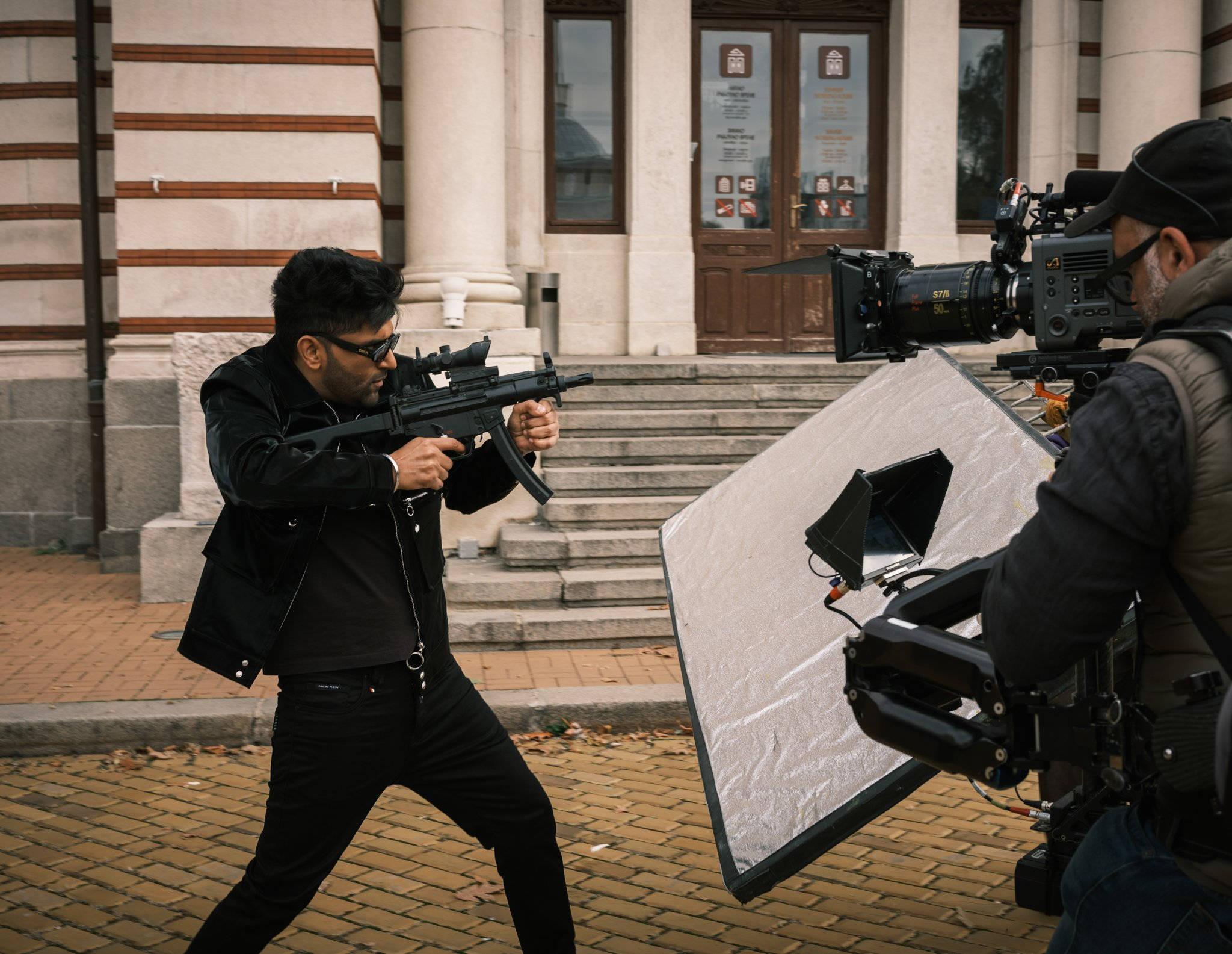 Guru Randhawa 桌布 壁紙 — 一名黑衣男子在磚街上瞄準步槍，附近有攝影師用大型反光板拍攝。