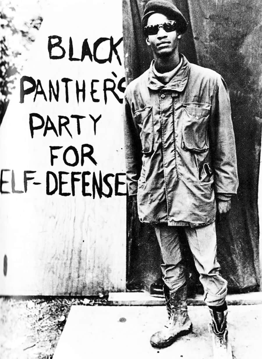 Black Panther Party 桌布 壁紙 — 一名黑豹黨成員自信地站在寫有「Black Panther's Party for Self-Defense」的標誌前，身處粗糙的城市環境中。