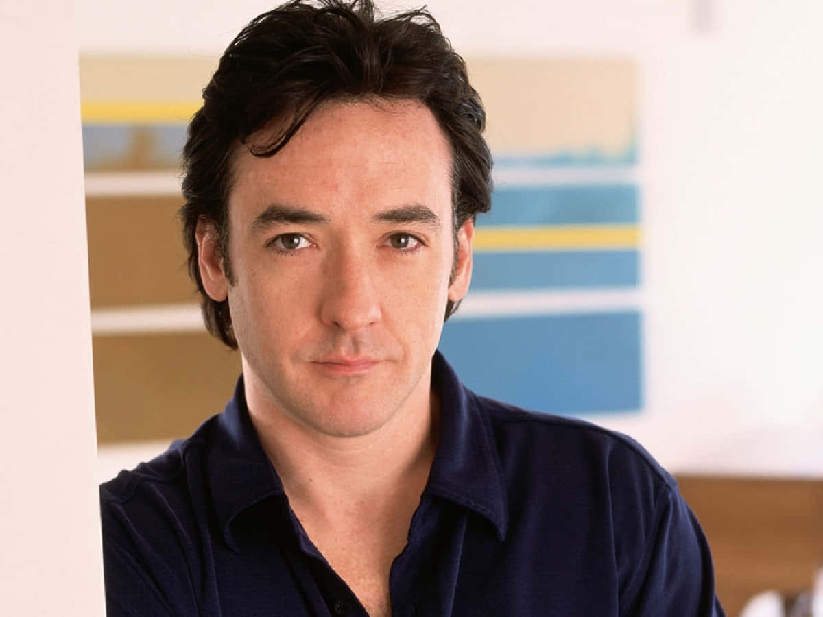 John Cusack 桌布 壁紙 — 一名黑髮、表情平靜的男子，身穿海軍藍襯衫，背景是色彩繽紛的抽象圖案。