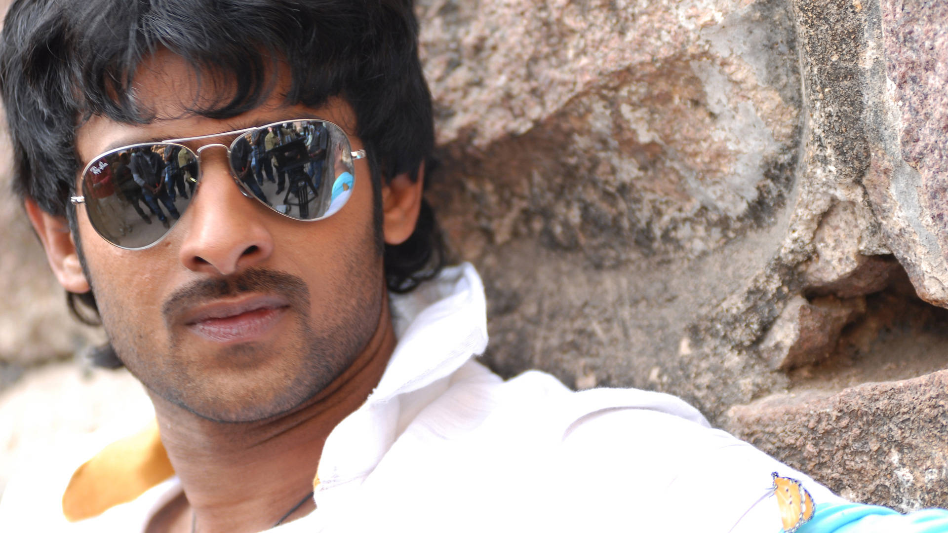 Prabhas HD 桌布 壁紙 — 一名黑髮戴太陽眼鏡的男子靠著粗糙的石牆，他的倒影在鏡片中顯示出一群人。